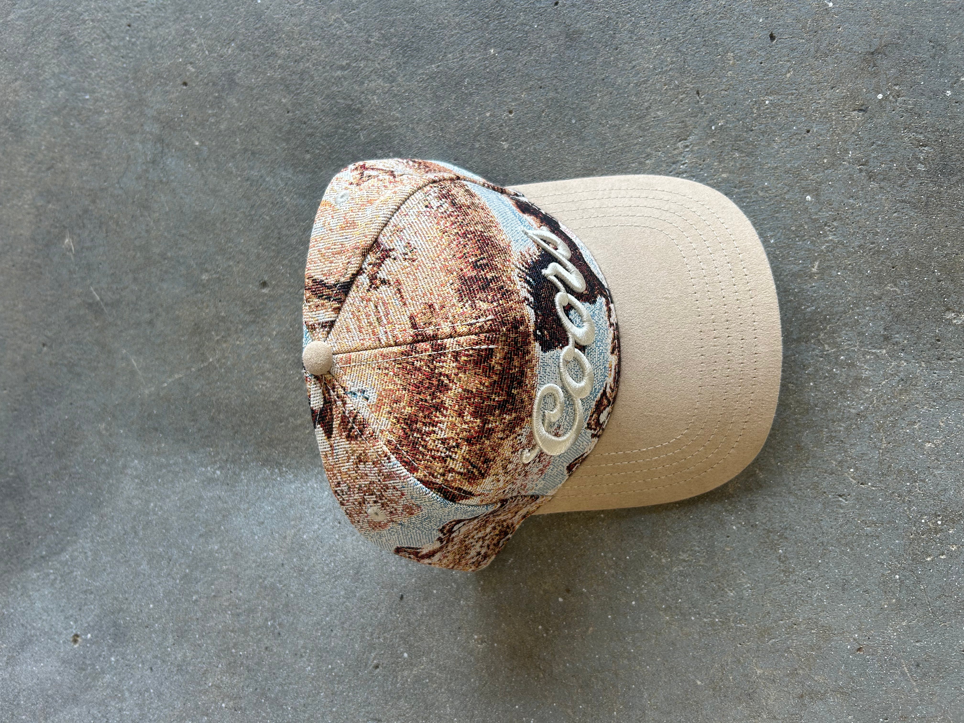 A baseball cap with a distressed brown and beige camouflage pattern on the front panels and a solid beige curved brim, featuring a white embroidered script logo on the side, displayed on a gray concrete surface