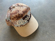 A Coors-branded baseball cap featuring a brown and beige camouflage pattern on the crown with a solid beige visor and embroidered Coors logo, resting on a gray concrete floor