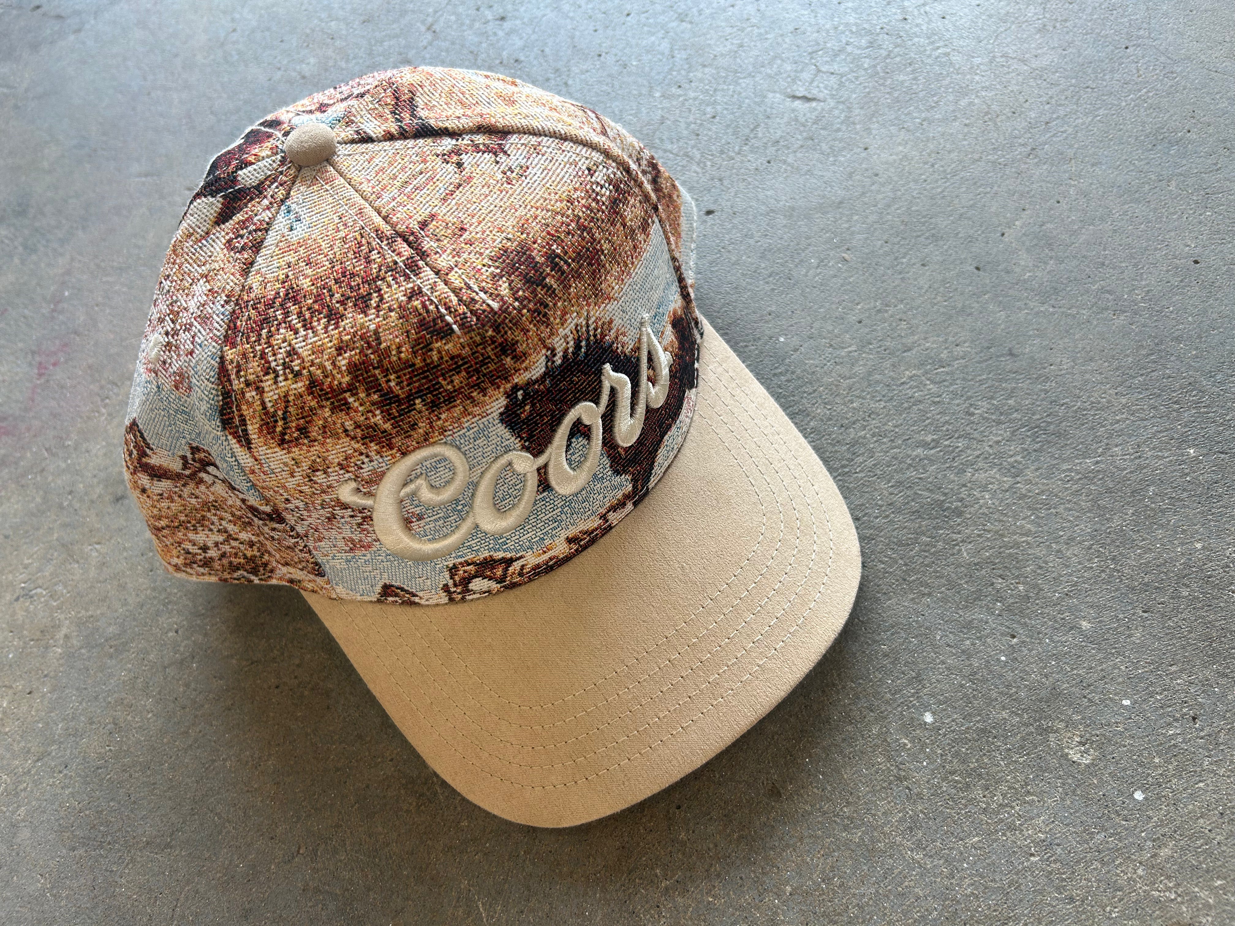 A Coors-branded baseball cap featuring a brown and beige camouflage pattern on the crown with a solid beige visor and embroidered Coors logo, resting on a gray concrete floor
