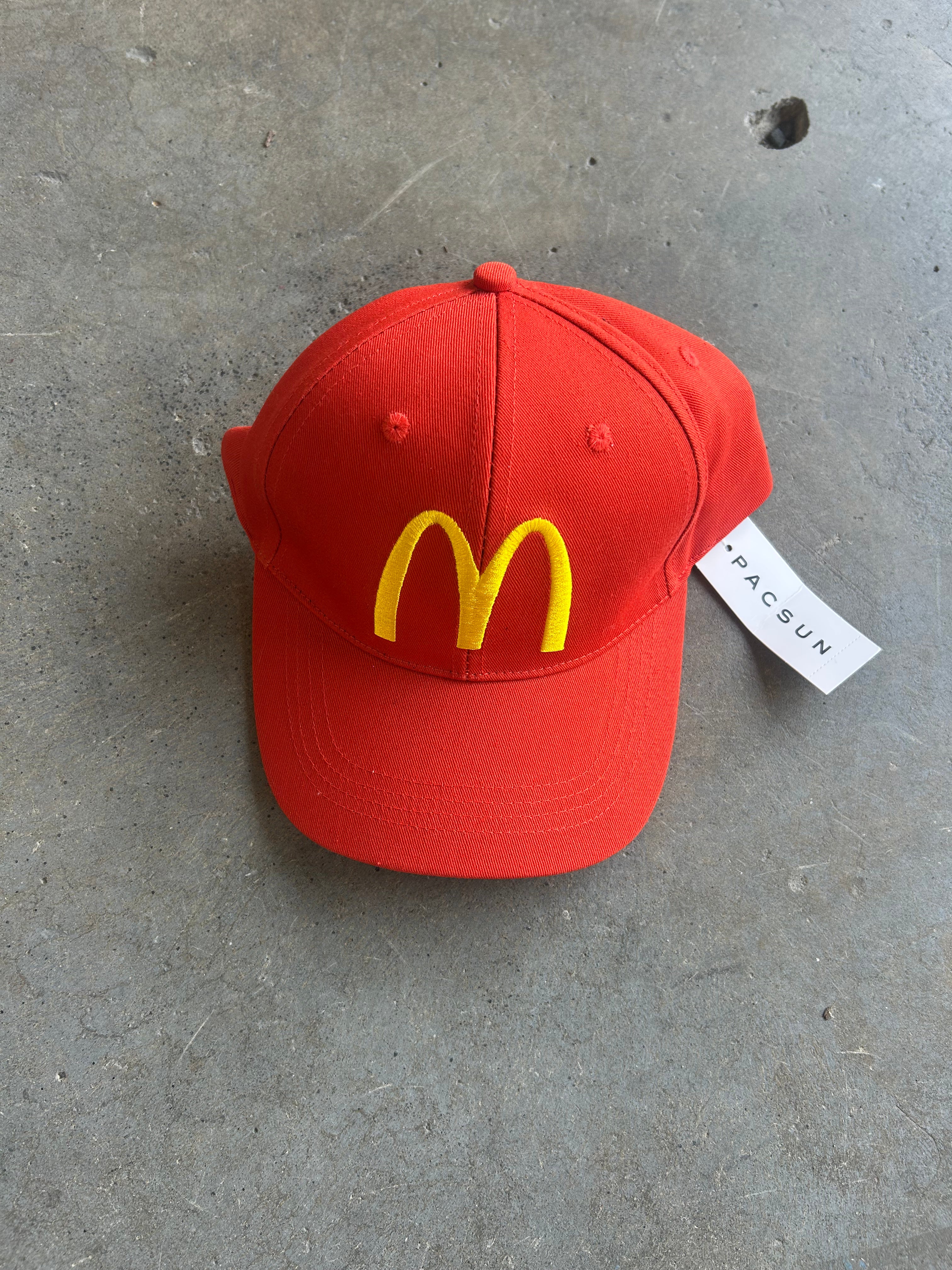 A red baseball cap featuring the yellow McDonald's arches logo embroidered on the front, resting on a gray concrete floor with a white 'PACSUN' brand tag attached
