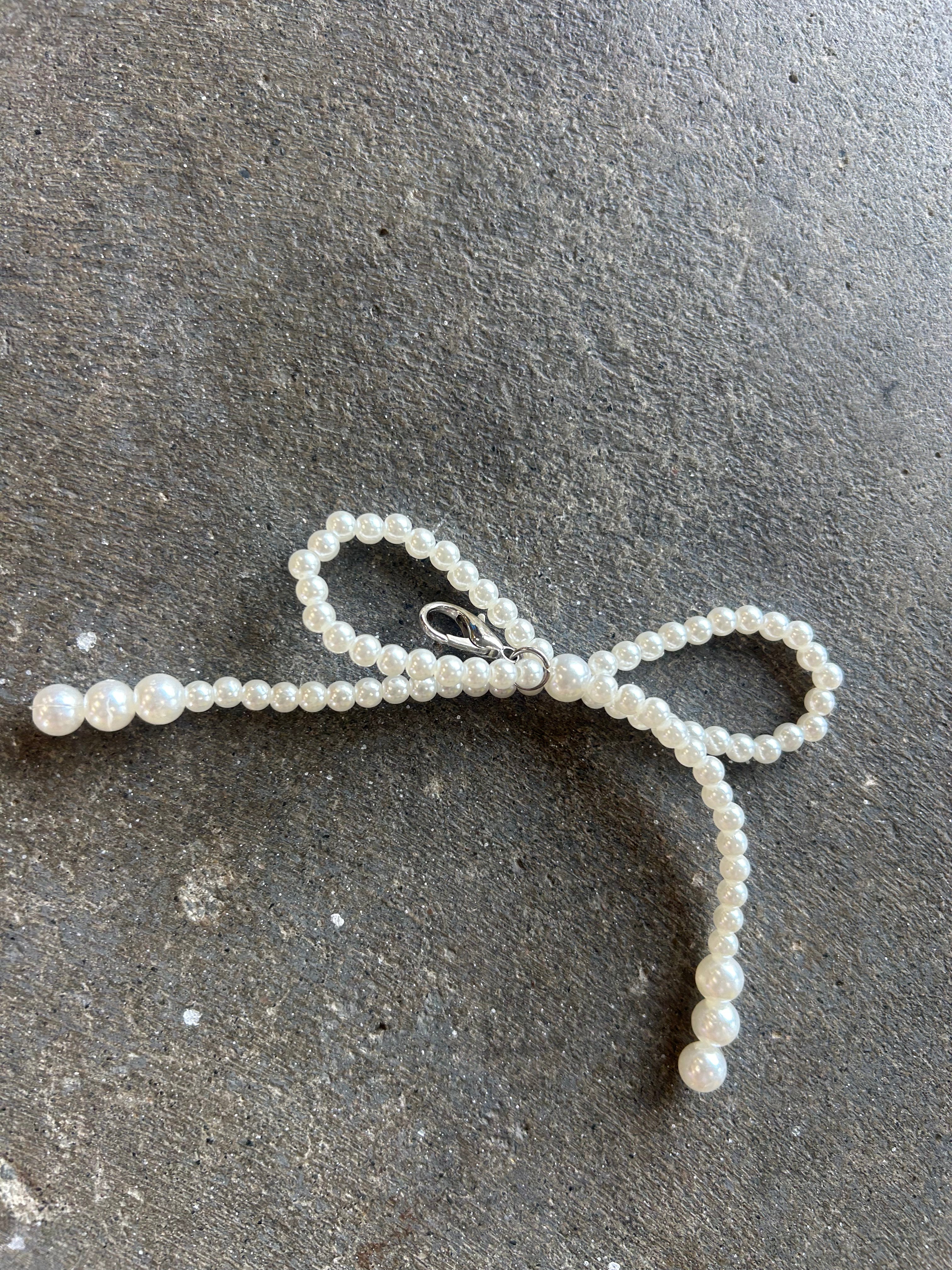 A white pearl beaded bow accessory with a silver lobster clasp, laid flat on a dark gray textured concrete surface.