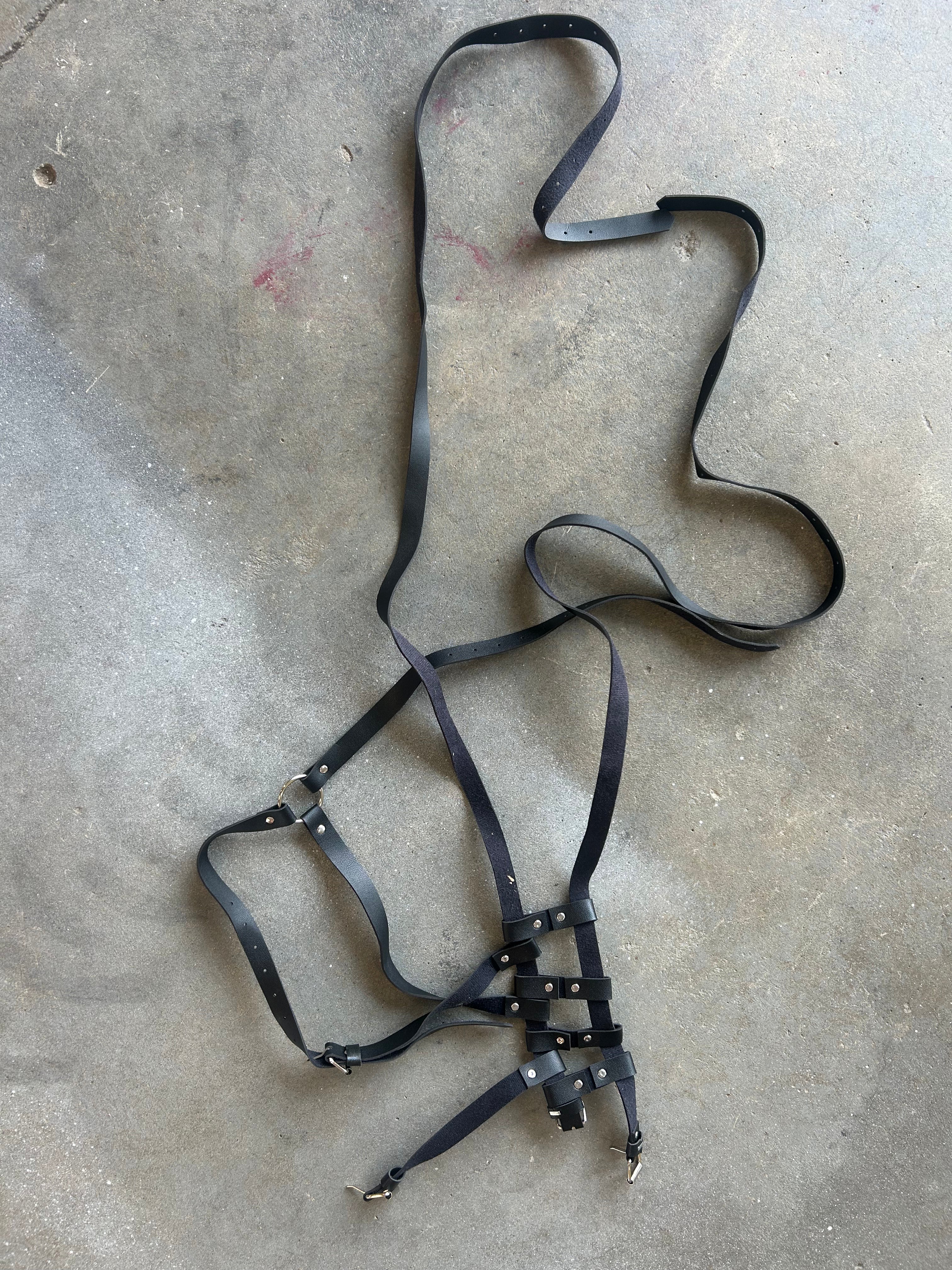 A black leather harness with adjustable straps and metal snap closures, laid out on a gray concrete floor