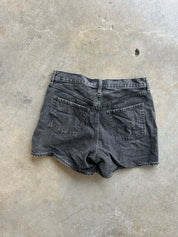 A pair of faded black denim shorts shown from the back, laid flat on a textured concrete floor