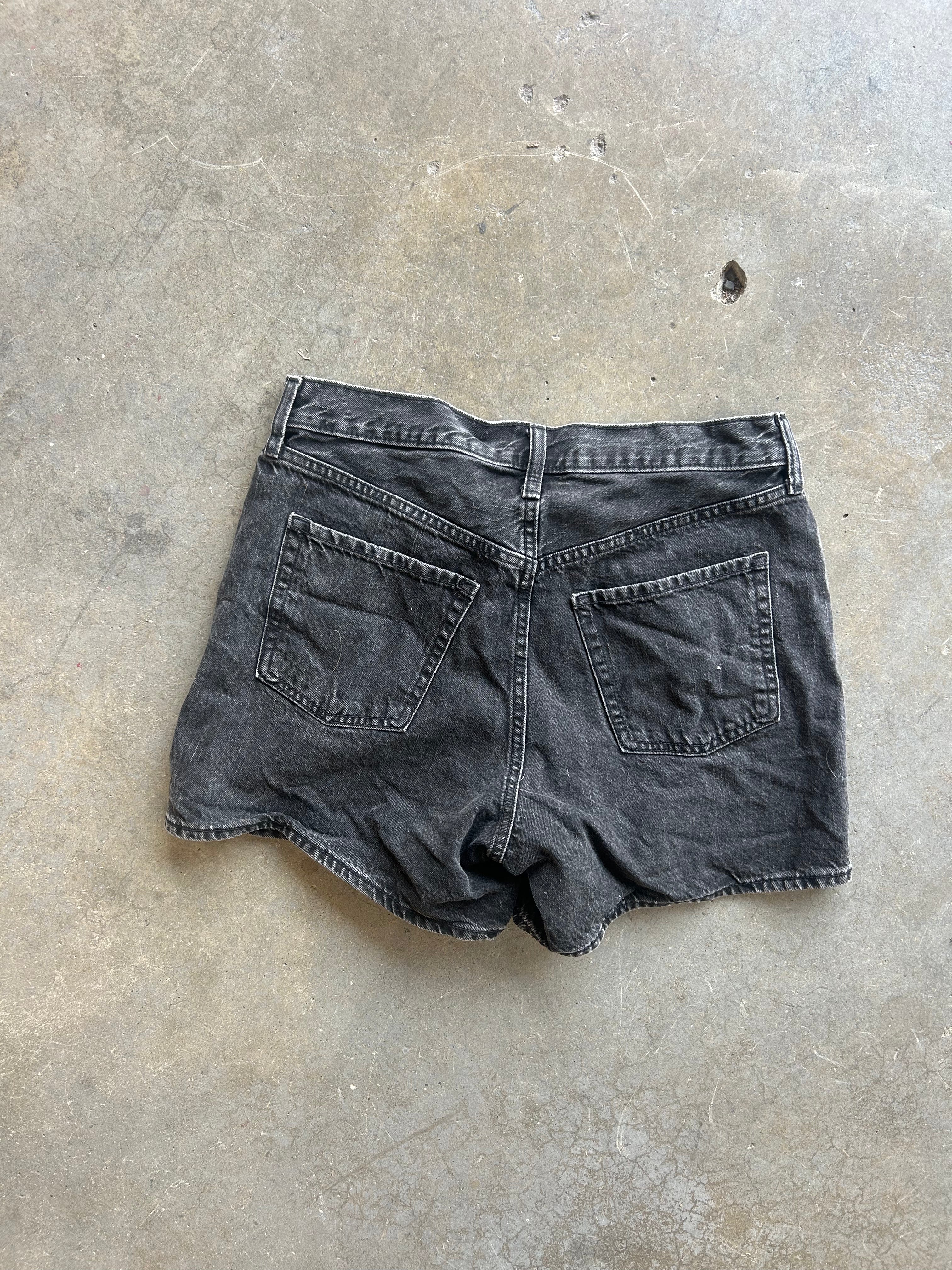 A pair of faded black denim shorts shown from the back, laid flat on a textured concrete floor
