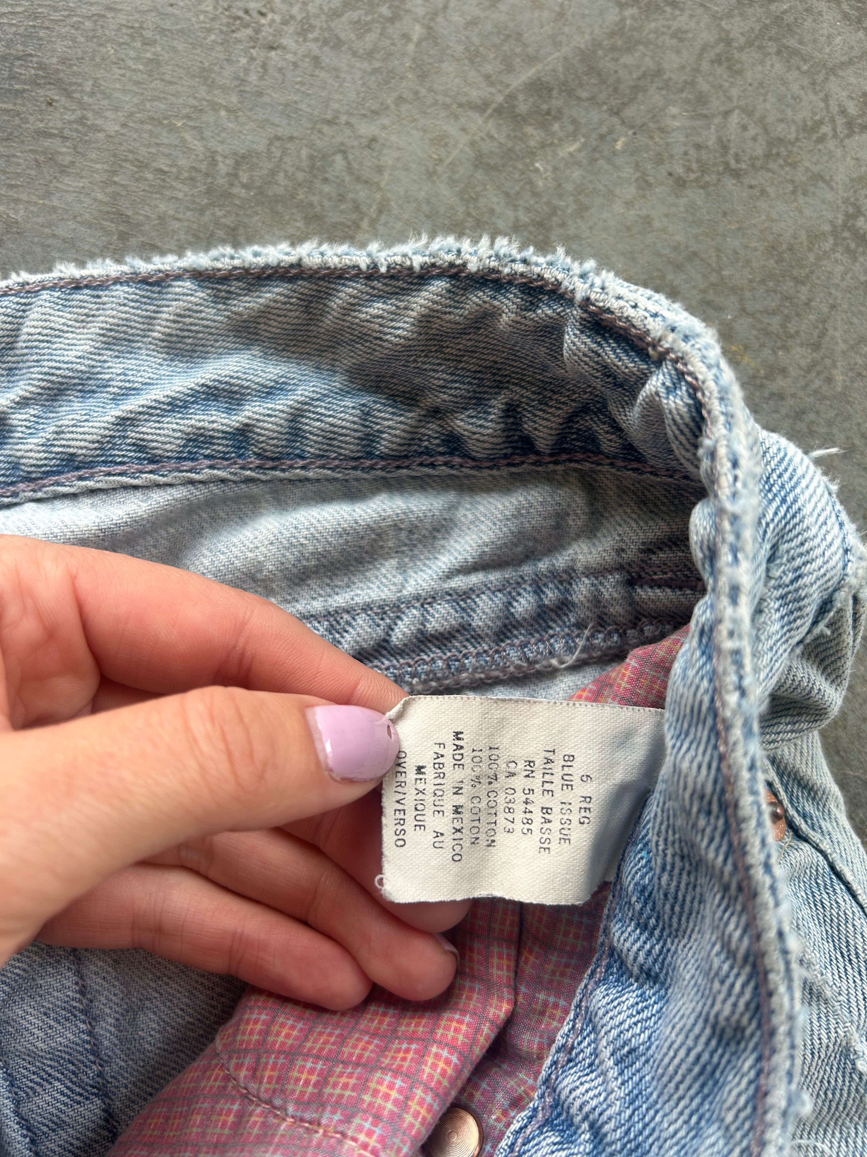 A close-up of a person's hand holding a white size and material label inside the waistband of light-wash blue denim jeans, showing a pink and red plaid lining
