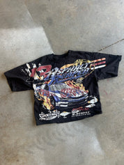 A black short-sleeve graphic T-shirt laid flat on a concrete floor, featuring a large front print of a race car with flames and the text '13 Ashland Speedway' and 'Blackout'