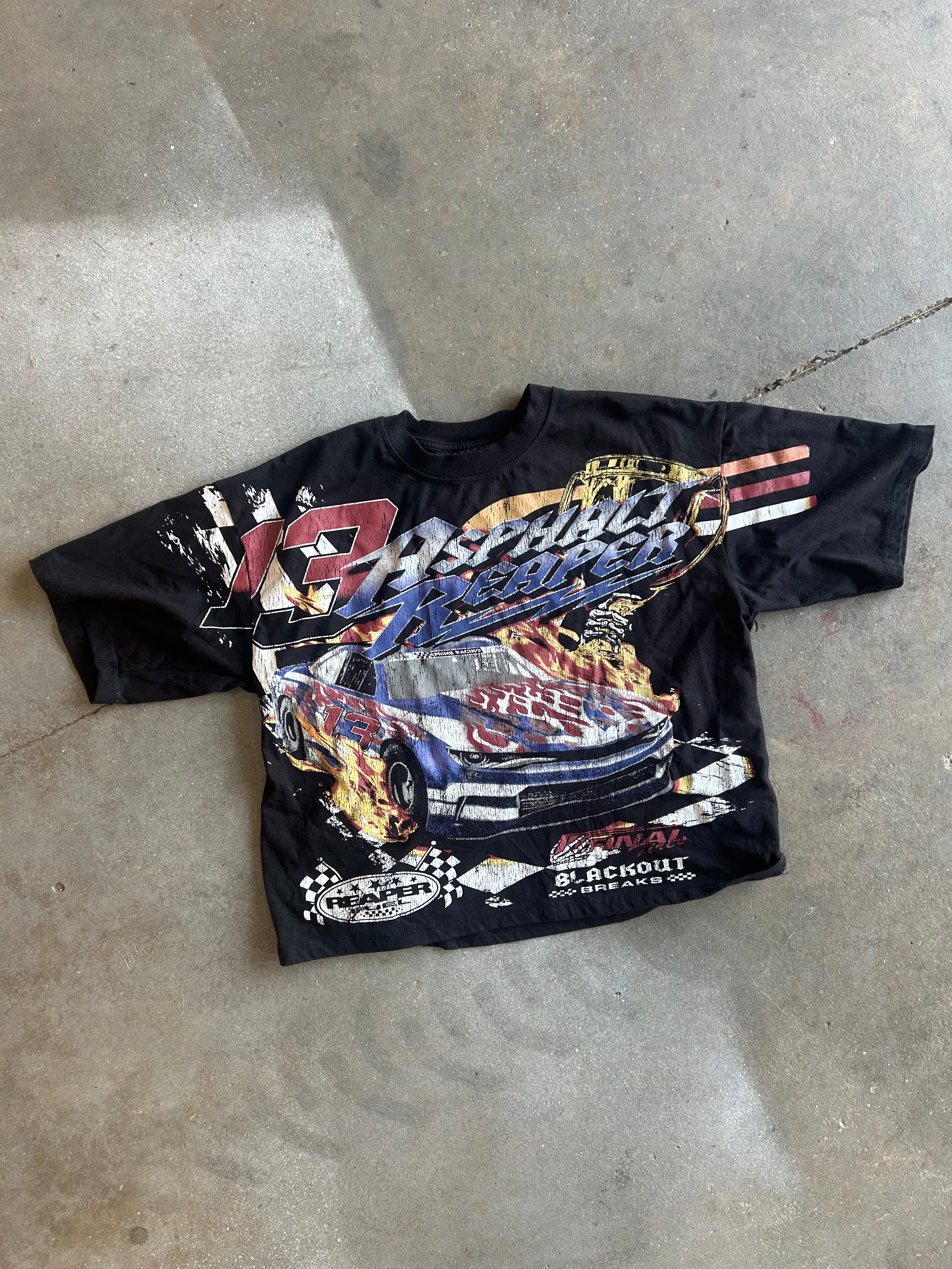 A black short-sleeve graphic T-shirt laid flat on a concrete floor, featuring a large front print of a race car with flames and the text '13 Ashland Speedway' and 'Blackout'