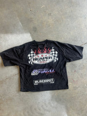 A black short-sleeve graphic T-shirt laid flat on a concrete floor. The shirt features a large front graphic with flames, checkered flags, the text 'Reaper Tool Box,' 'Final 14,' and 'Blackout