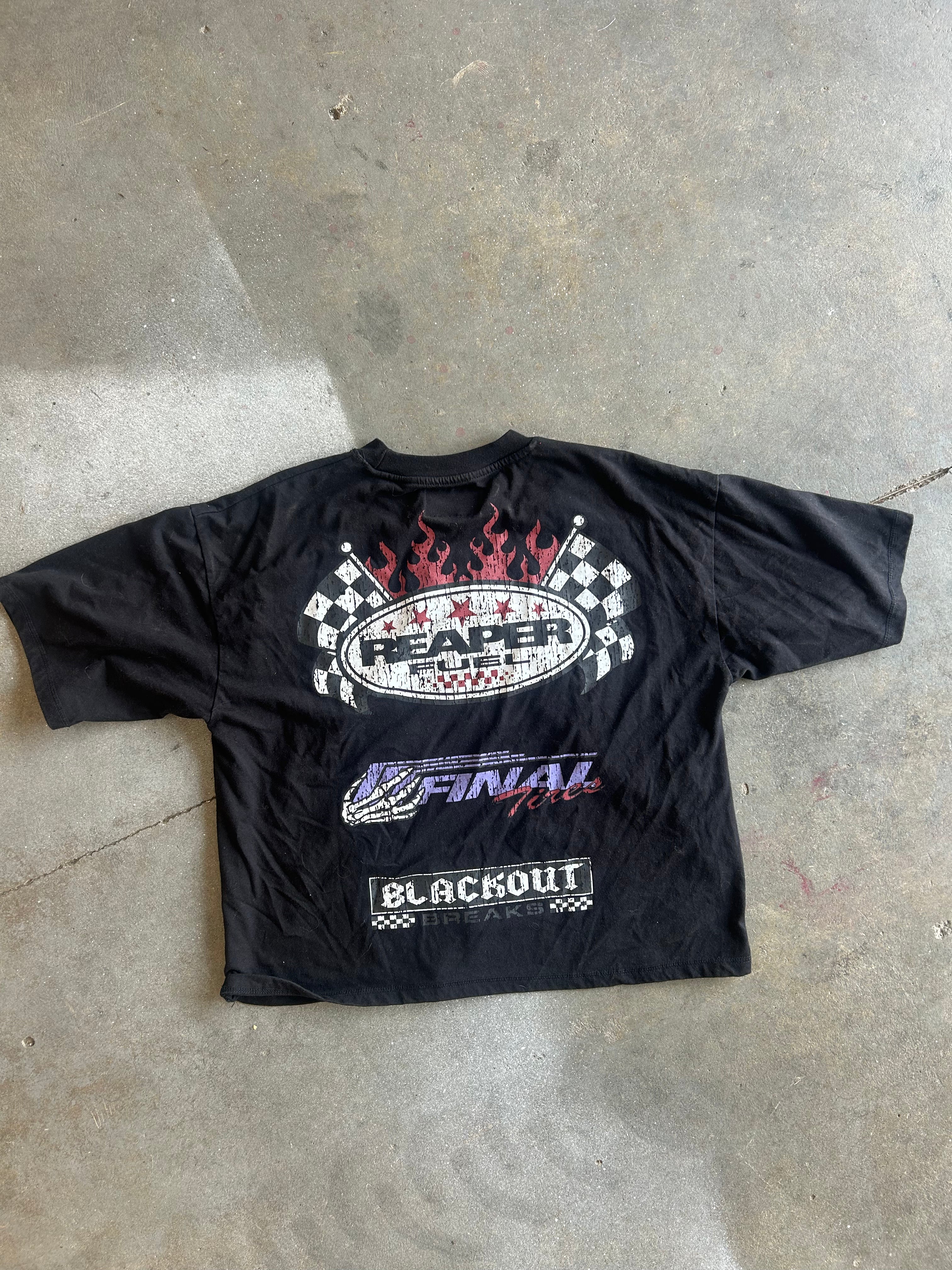 A black short-sleeve graphic T-shirt laid flat on a concrete floor. The shirt features a large front graphic with flames, checkered flags, the text 'Reaper Tool Box,' 'Final 14,' and 'Blackout