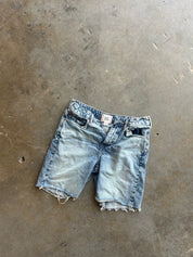 Light wash denim shorts with a raw, frayed hem, laid flat on a textured concrete floor