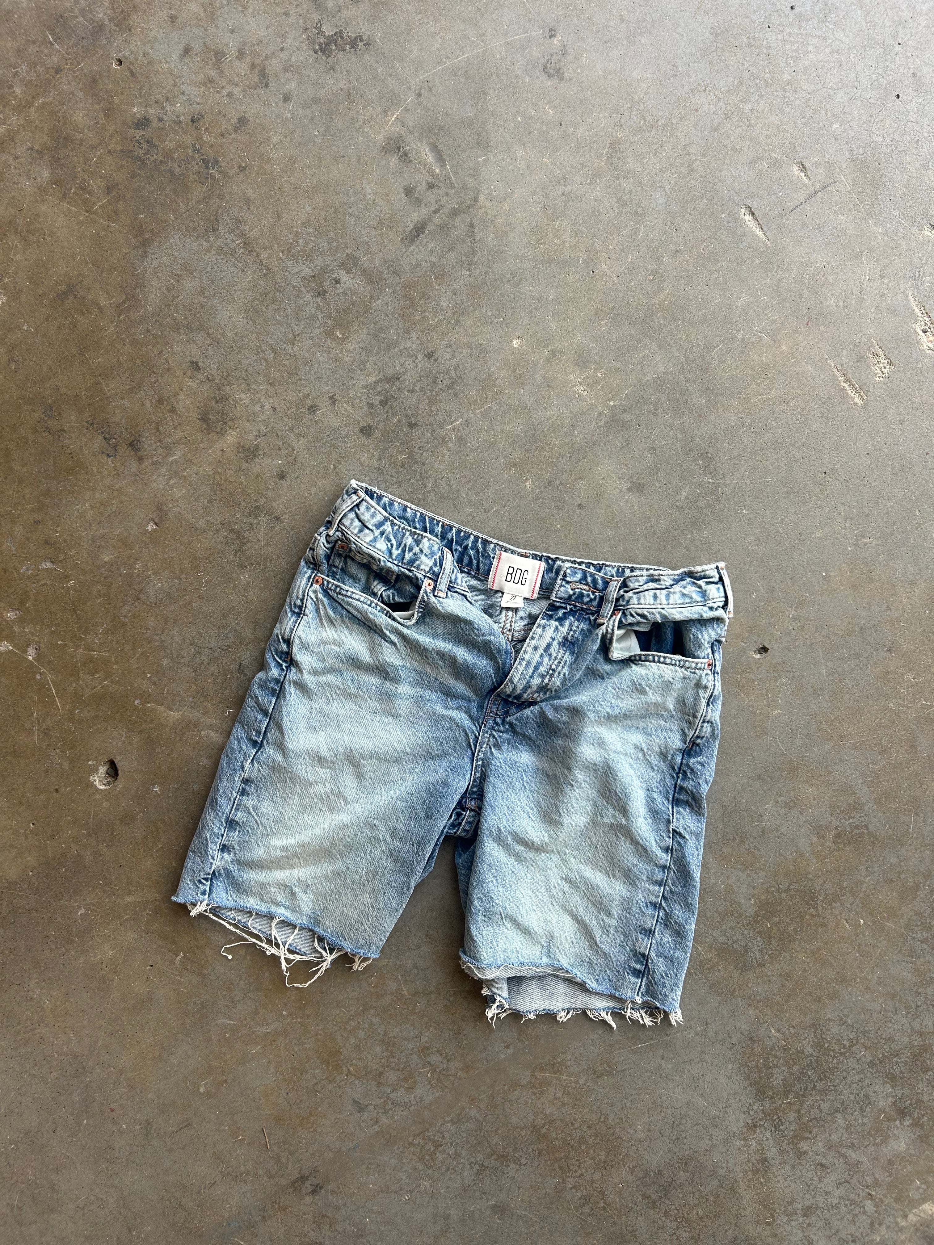 Light wash denim shorts with a raw, frayed hem, laid flat on a textured concrete floor