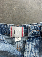Close-up of the waistband of blue denim jeans with a white BDG brand label stitched onto the back waistband, set against a gray concrete background