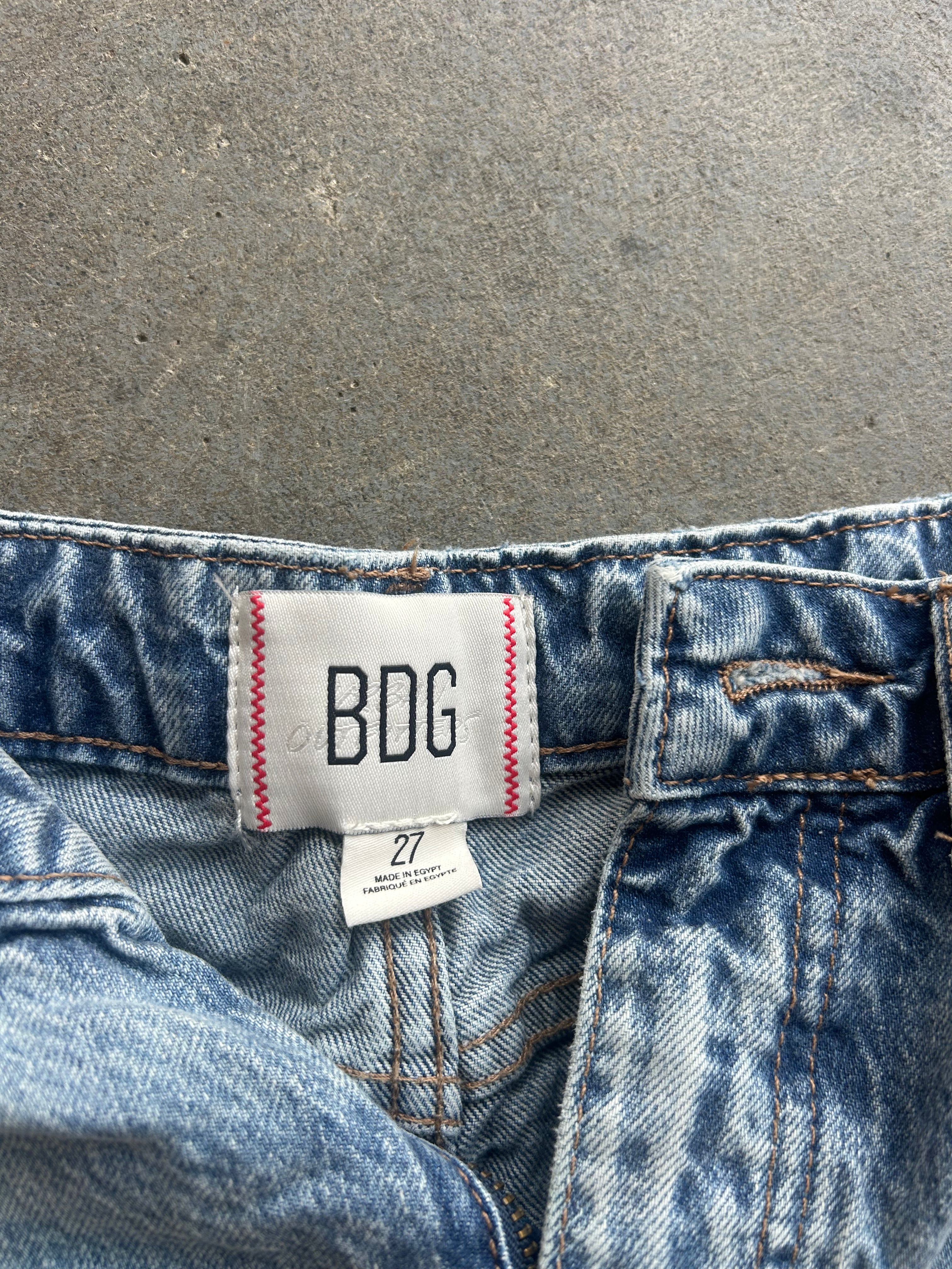 Close-up of the waistband of blue denim jeans with a white BDG brand label stitched onto the back waistband, set against a gray concrete background