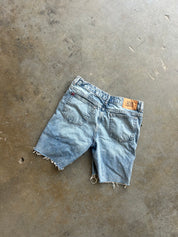 Light wash denim shorts with raw frayed hems, laid flat on a textured concrete floor. The back pocket and a small orange leather brand patch are visible