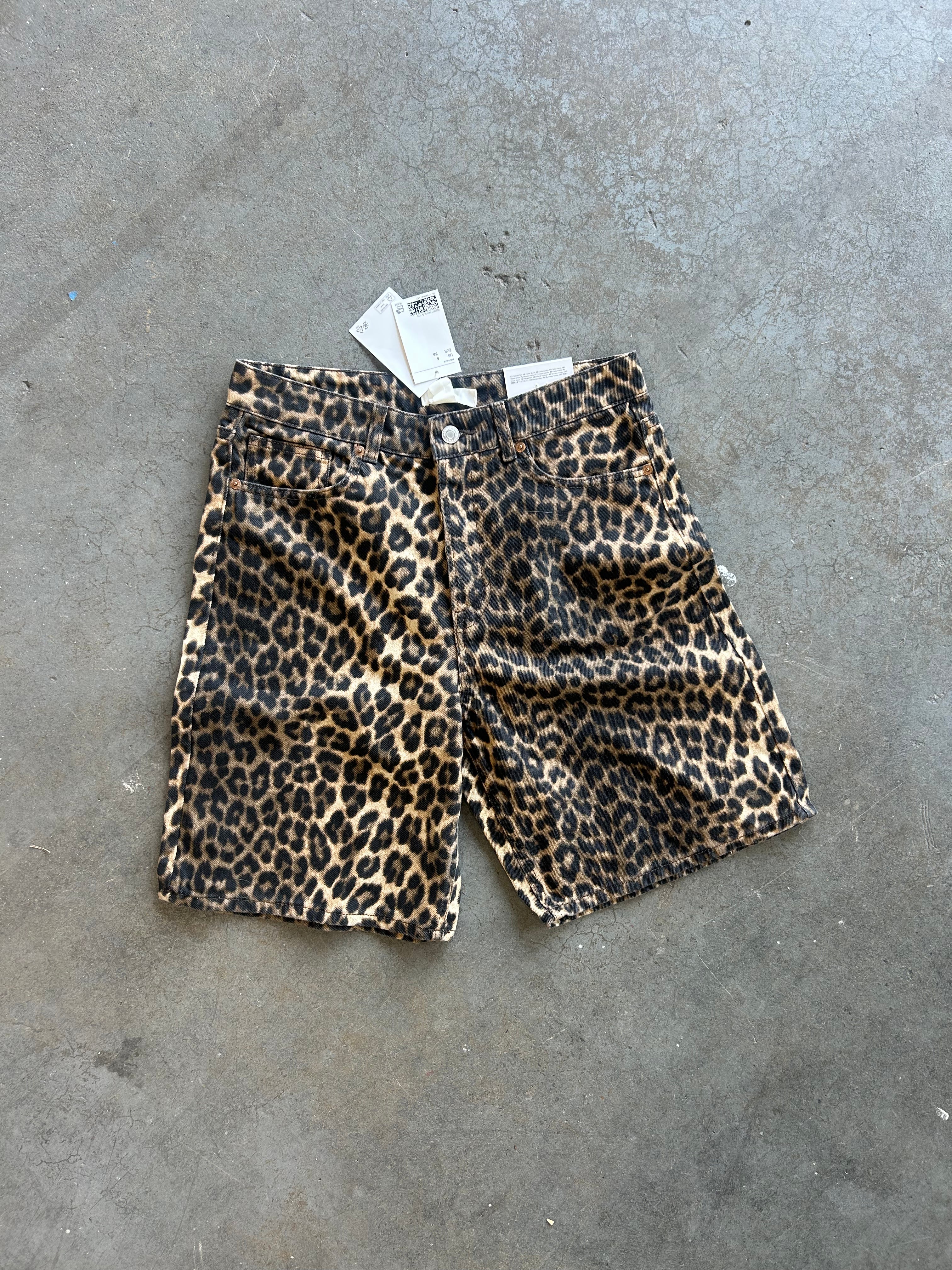 A pair of leopard print shorts with a white price tag attached, laid flat on a gray concrete floor