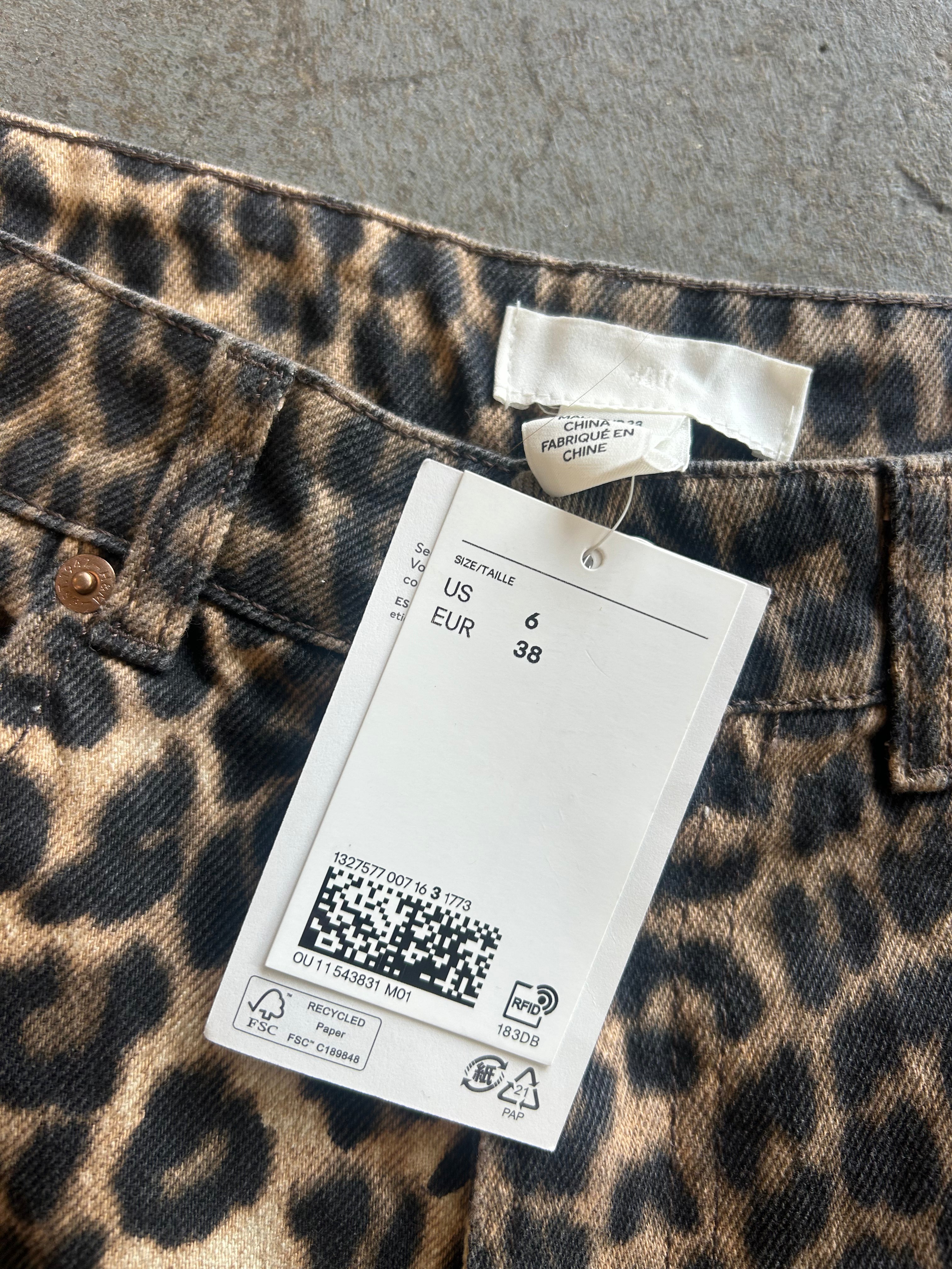 Close-up of leopard-print jeans with a white size tag attached, showing the waistband, belt loops, and fabric pattern on a gray surface