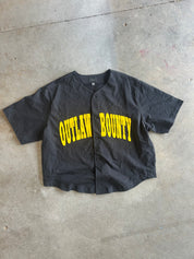 A black short-sleeve button-up baseball-style shirt with 'OUTLAW BOUNTY' printed across the front in bold yellow letters, laid flat on a concrete floor