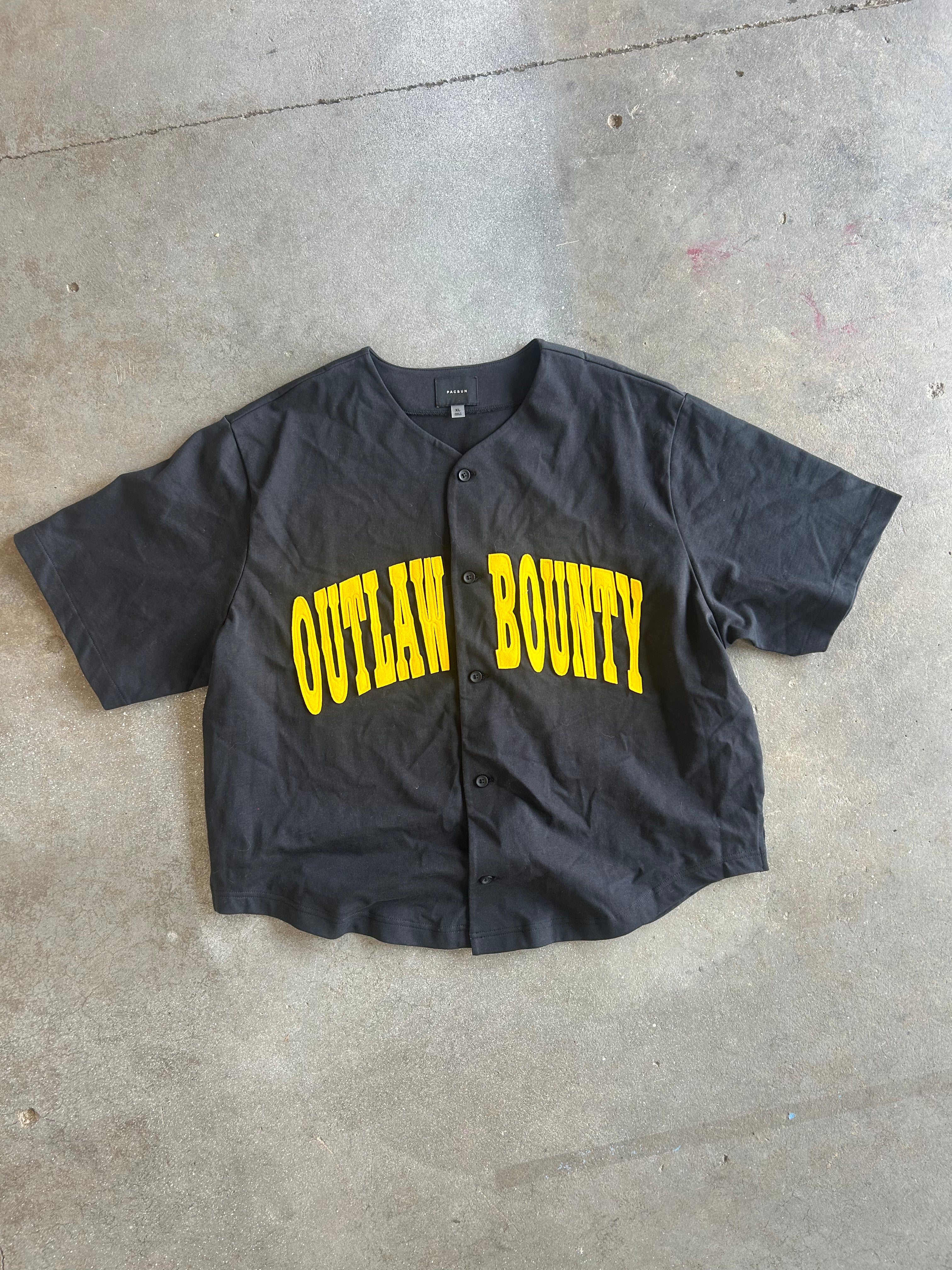 A black short-sleeve button-up baseball-style shirt with 'OUTLAW BOUNTY' printed across the front in bold yellow letters, laid flat on a concrete floor
