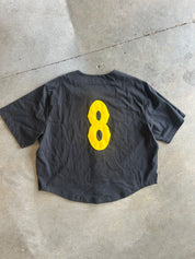A black short-sleeve t-shirt laid flat on a concrete floor, featuring a large yellow number 8 printed on the back