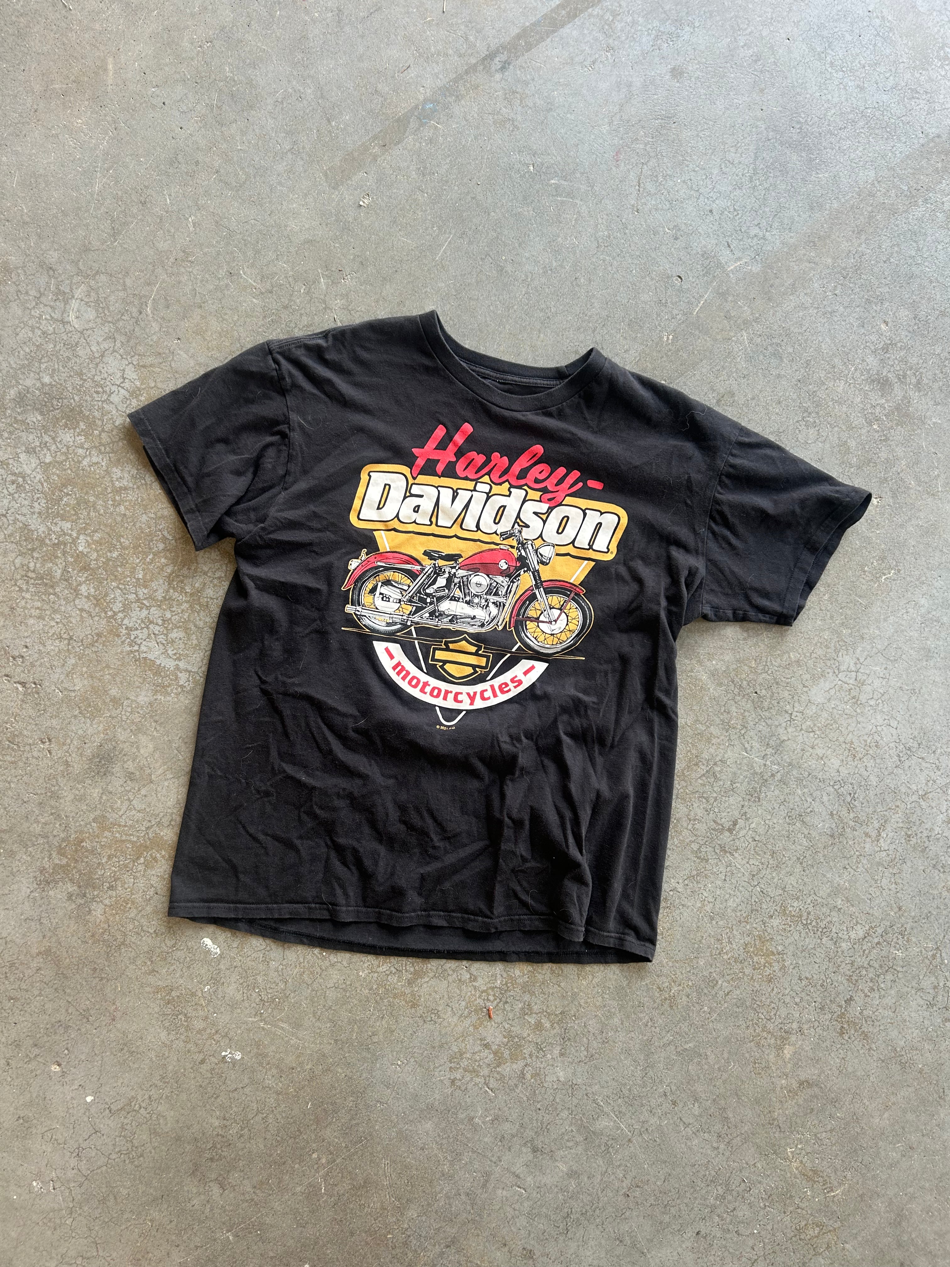 A black vintage Harley-Davidson graphic t-shirt laid flat on a concrete floor. The shirt features a large front print with the words 'Harley-Davidson' in red and yellow, along with a motorcycle illustration 