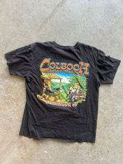 A black vintage graphic t-shirt laid flat on a concrete floor. The shirt features a large front graphic with the text 'COLBOCH HARLEY-DAVIDSON' in orange and yellow, above a colorful illustration of a person riding a motorcycle in a green outdoor setting, with 'MERRISTOWN, TN' printed below