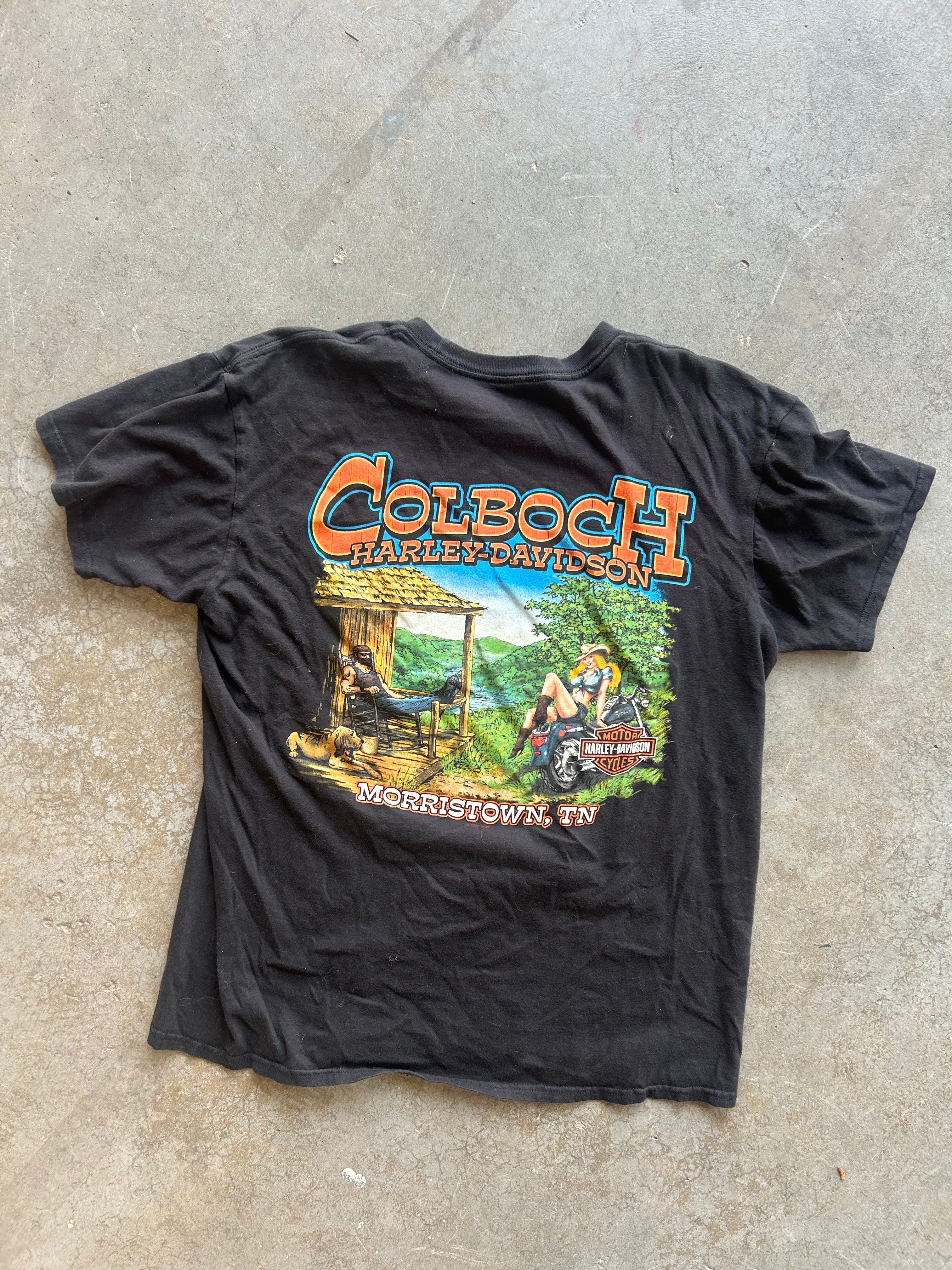 A black vintage graphic t-shirt laid flat on a concrete floor. The shirt features a large front graphic with the text 'COLBOCH HARLEY-DAVIDSON' in orange and yellow, above a colorful illustration of a person riding a motorcycle in a green outdoor setting, with 'MERRISTOWN, TN' printed below