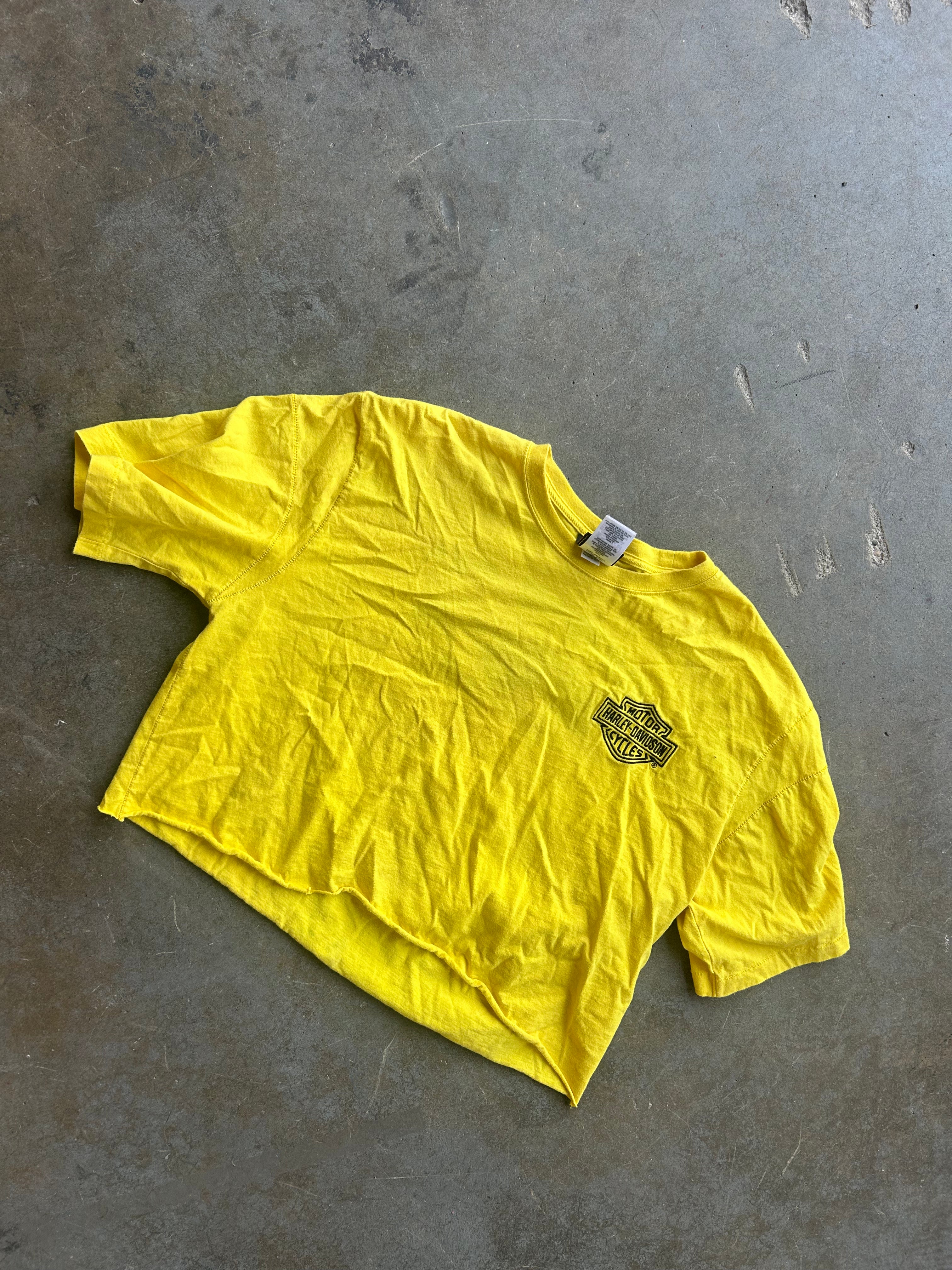 A bright yellow short-sleeve t-shirt with a small black graphic on the left chest, laid flat on a textured gray concrete floor