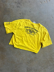 A bright yellow cropped short-sleeve T-shirt laid flat on a gray concrete floor, featuring a black Harley-Davidson winged logo and text printed on the back