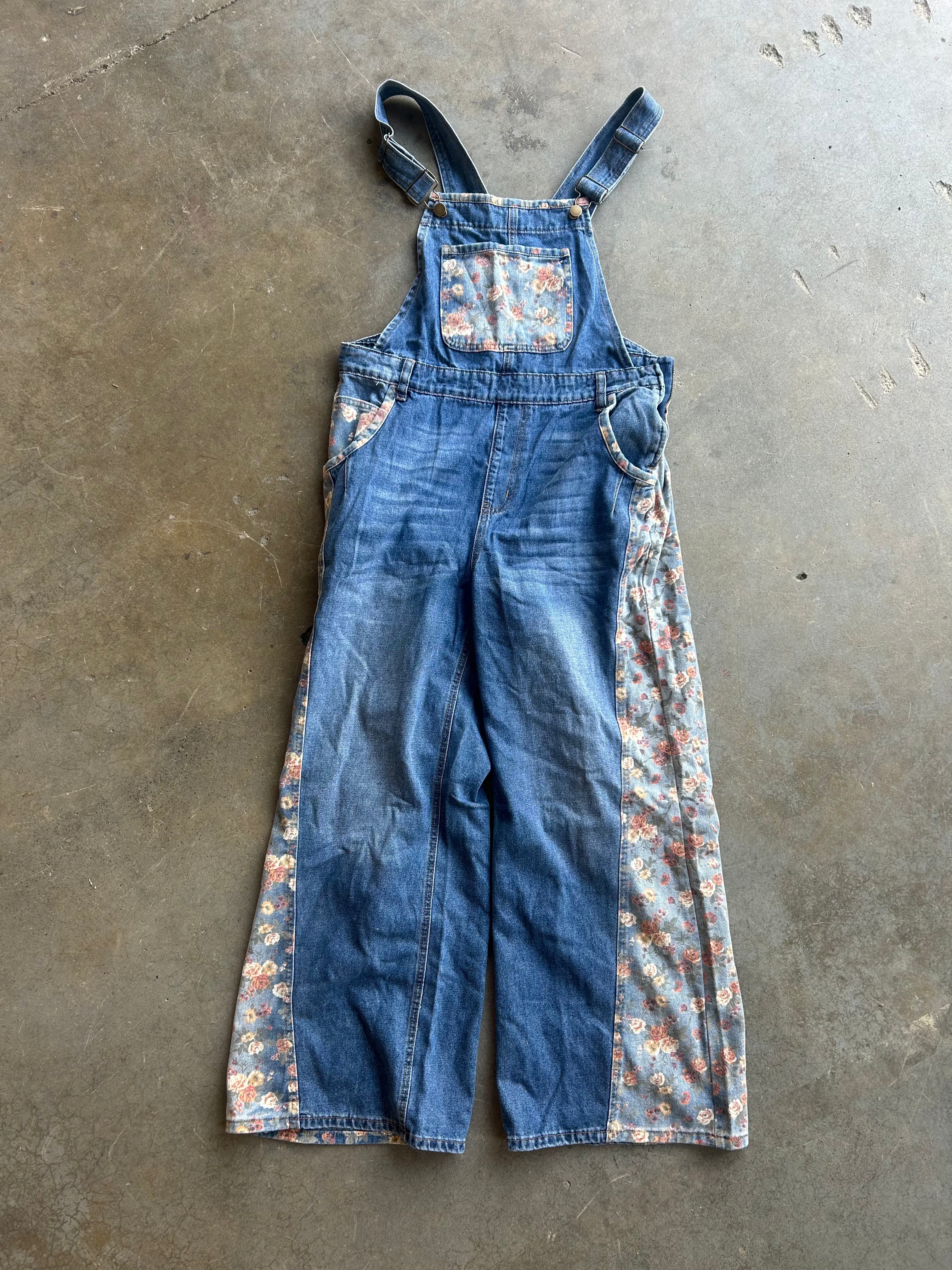 A pair of blue denim bib overalls with floral patterned side panels and adjustable shoulder straps, laid flat on a concrete floor