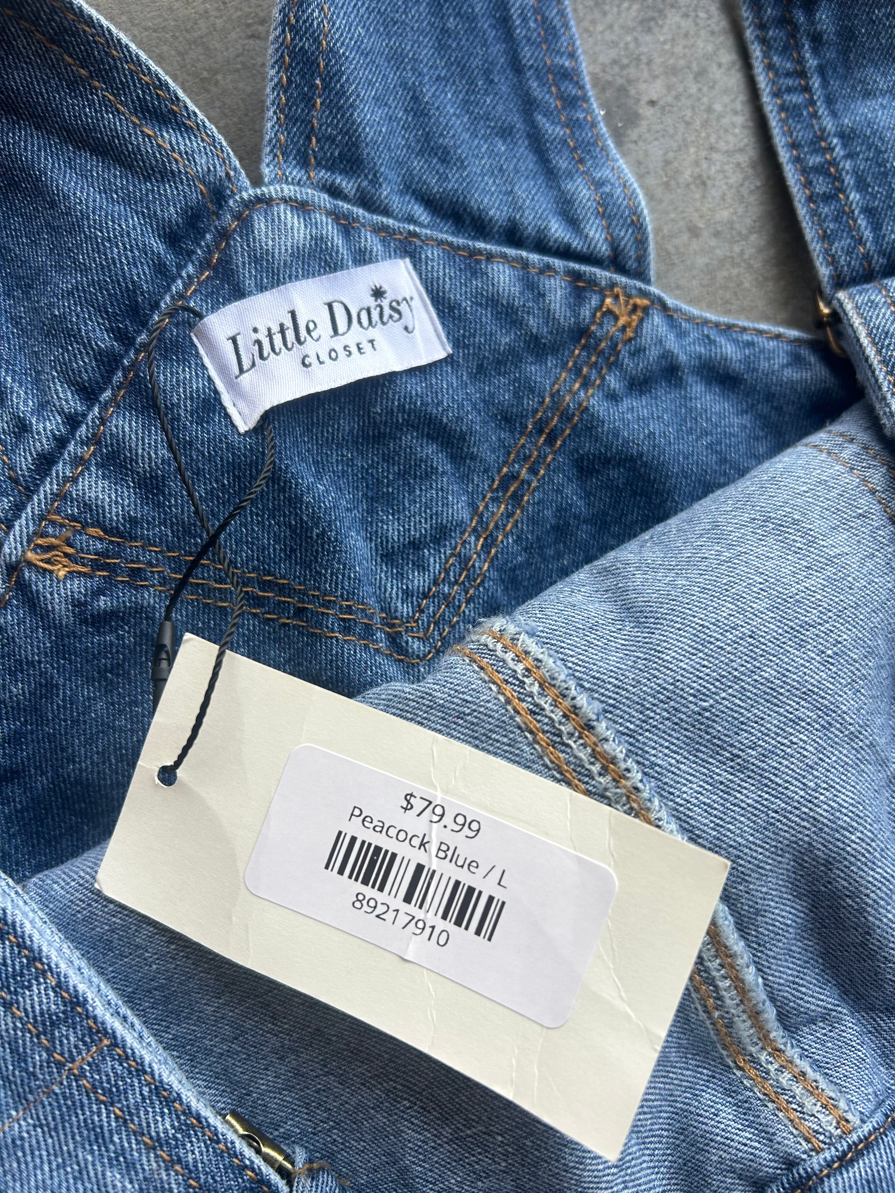 Close-up of a pair of blue denim overalls with a visible 'Little Daisy Closet' brand label and a price tag reading '$79.99 Peacock Blue / L'
