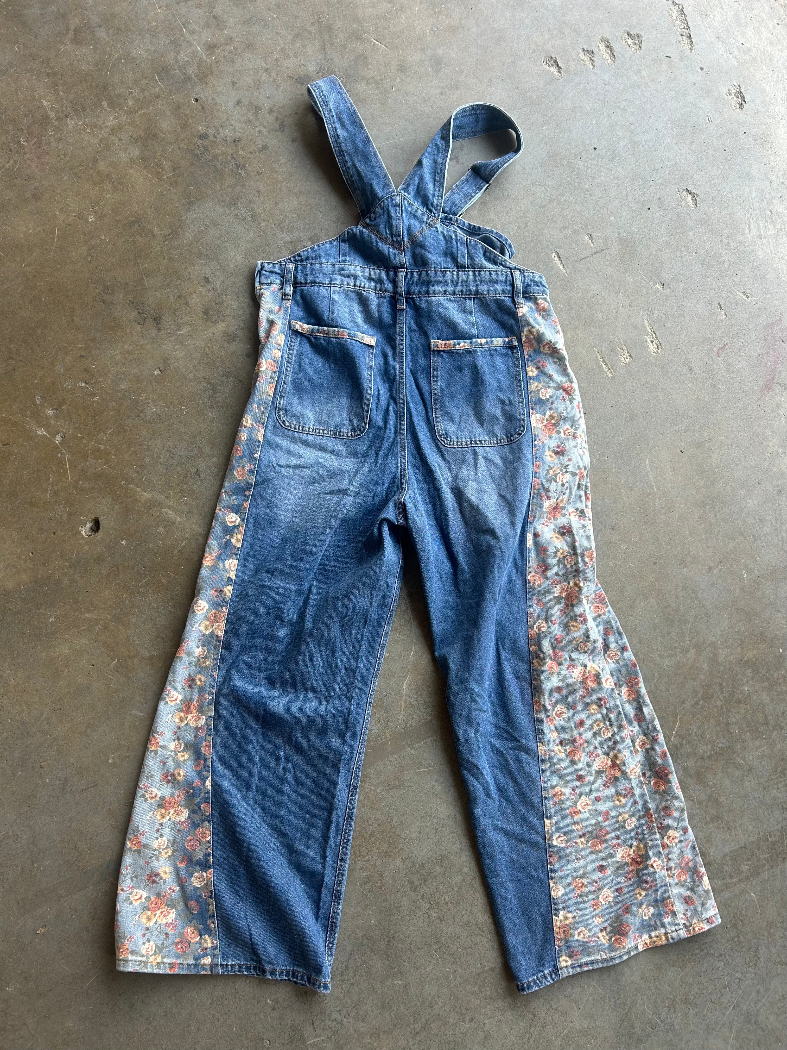 A pair of vintage-style denim bib overalls laid flat on a concrete floor, featuring blue denim on the front and floral patterned side panels in shades of pink, orange, and green