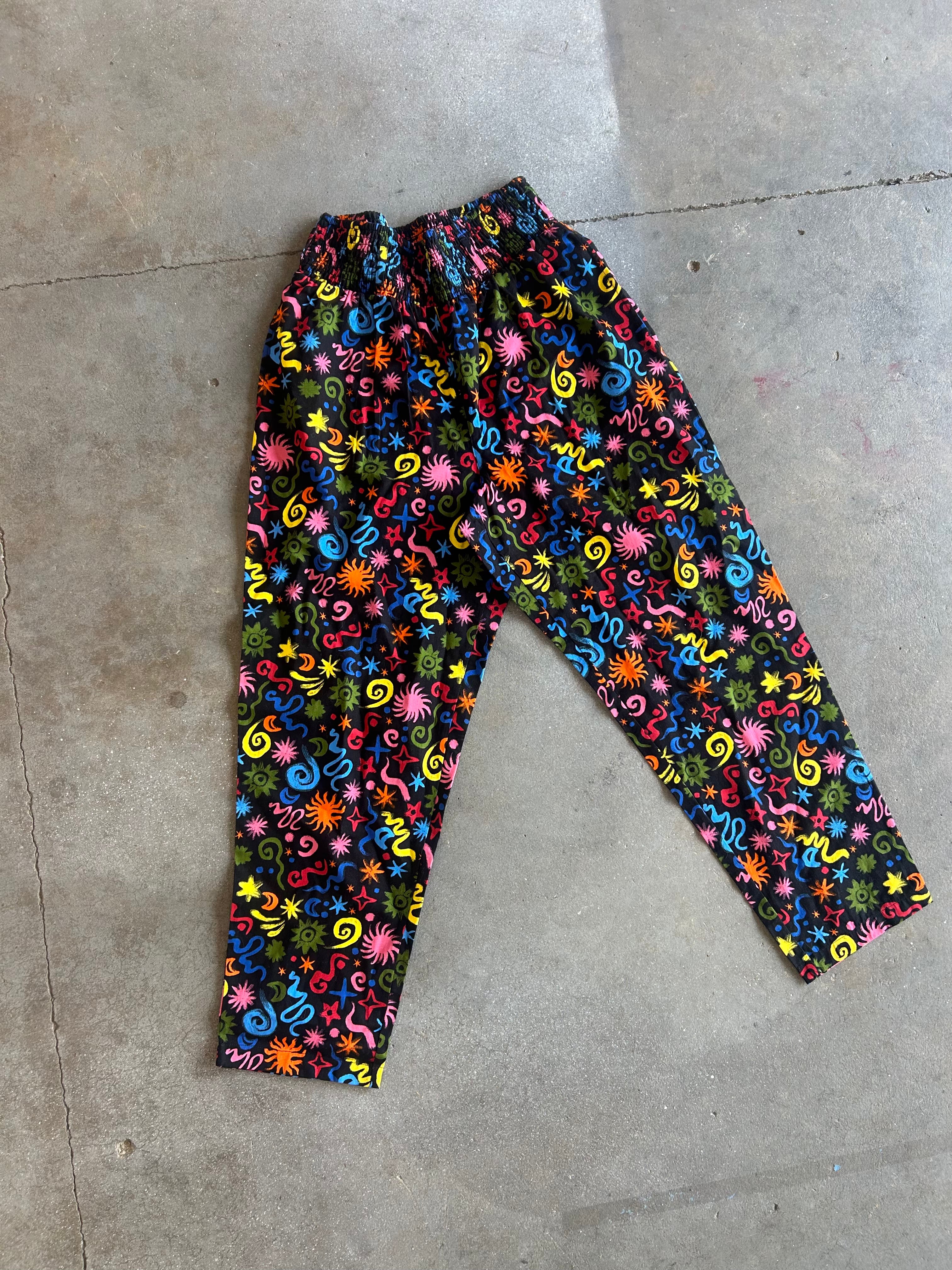 A pair of pants with a vibrant, multicolored floral and swirl pattern on a black background, laid flat on a gray concrete floor