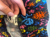 A close-up of a colorful woven textile with a hand holding a white fabric tag that reads 'Tula' and 'M/F'. The fabric has a black base with vibrant multicolor floral and geometric patterns in blue, pink, orange, yellow, and green