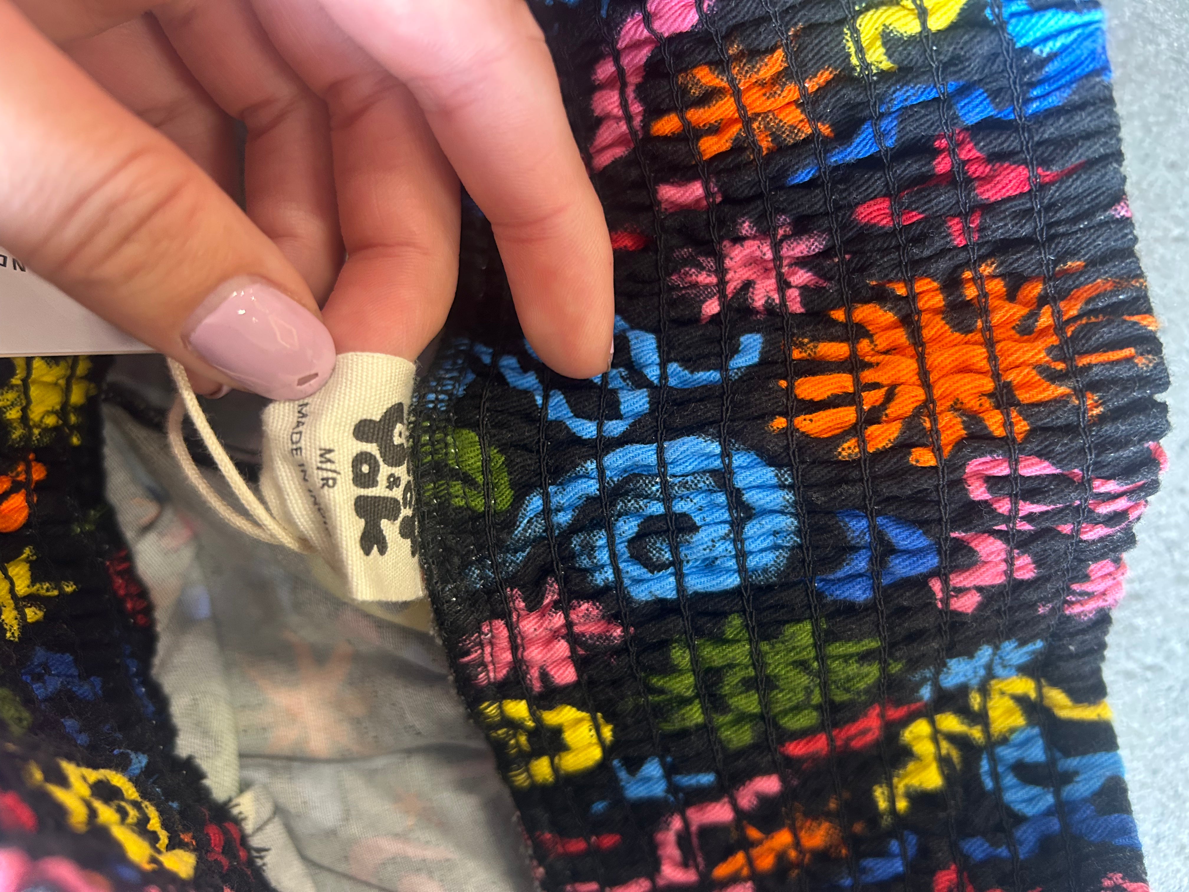 A close-up of a colorful woven textile with a hand holding a white fabric tag that reads 'Tula' and 'M/F'. The fabric has a black base with vibrant multicolor floral and geometric patterns in blue, pink, orange, yellow, and green