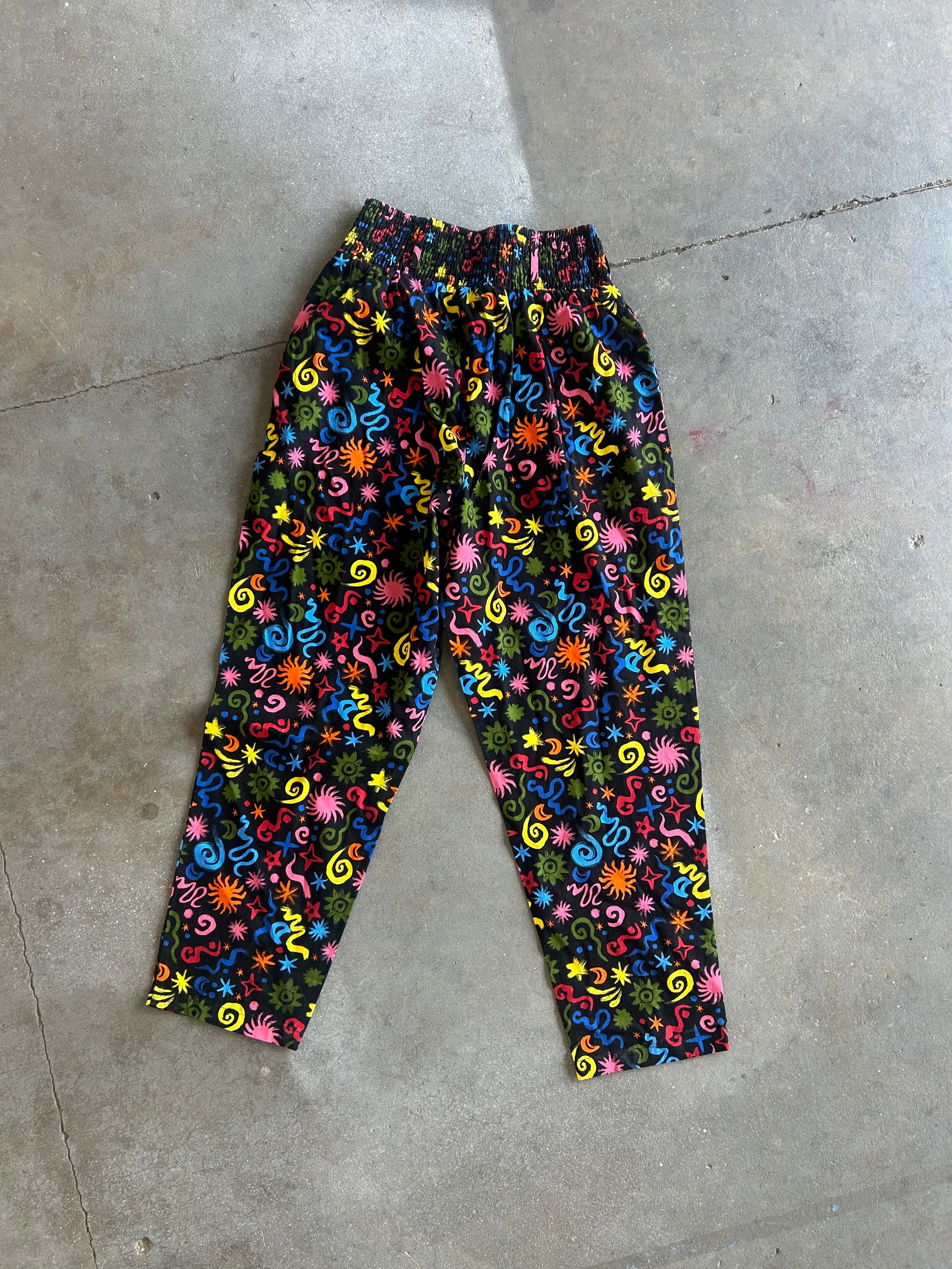 A pair of cropped pants with a vibrant, multicolored psychedelic swirl and floral pattern on a black background, laid flat on a gray concrete floor