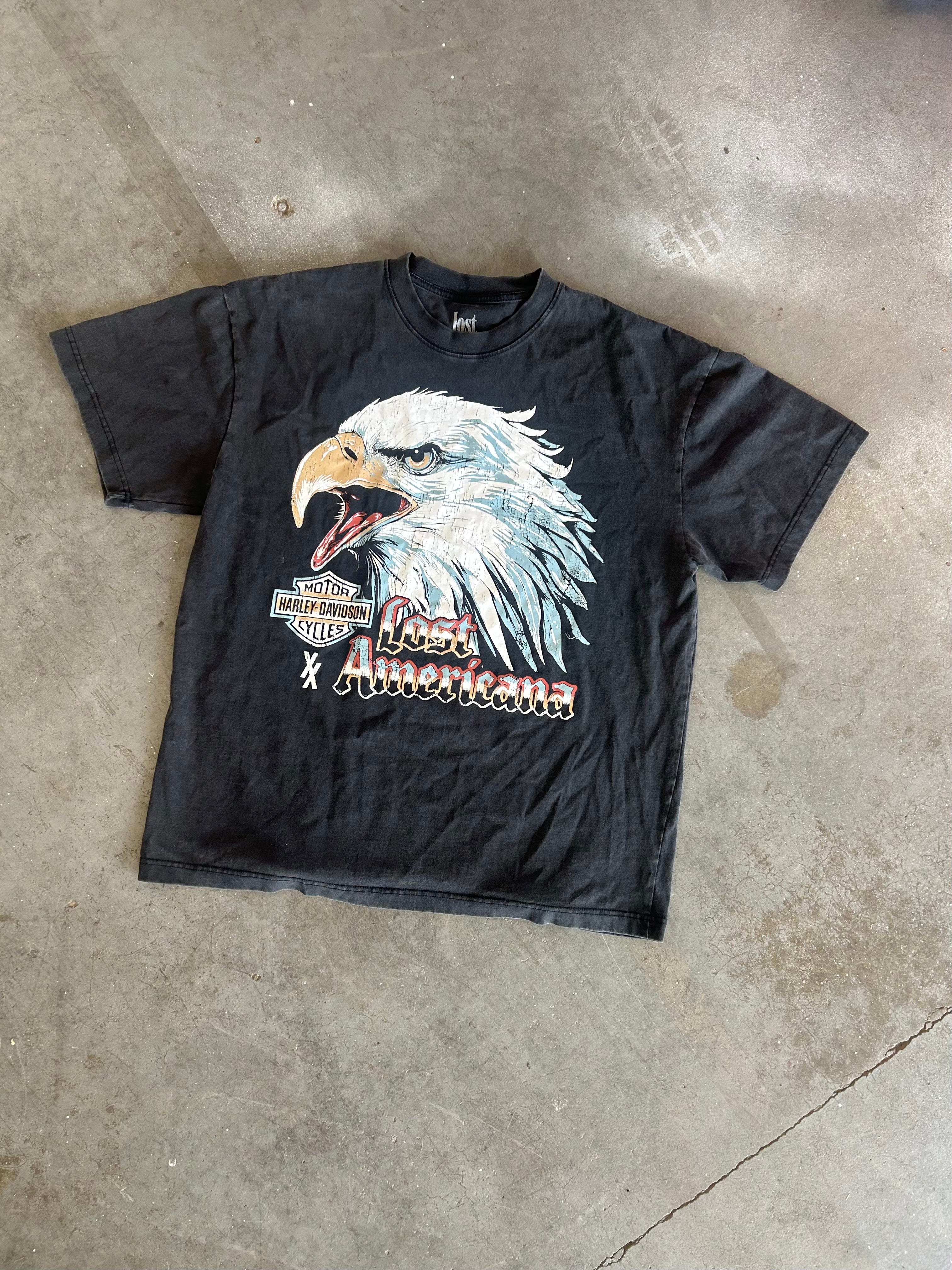 A faded black short-sleeve graphic t-shirt featuring a large bald eagle print and the text 'Lost Americans' with a motorcycle logo, laid flat on a concrete floor