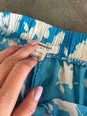A close-up of a person's hand with pink manicured nails pointing to the brand label of a blue and white patterned garment. The elastic waistband and brand name
