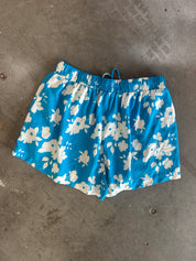 Blue and white floral print athletic shorts with an elastic waistband and drawstring, laid flat on a gray concrete floor