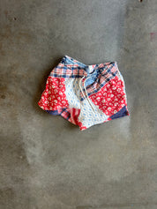 A pair of patchwork shorts with red, white, and blue patterns, laid flat on a gray concrete surface