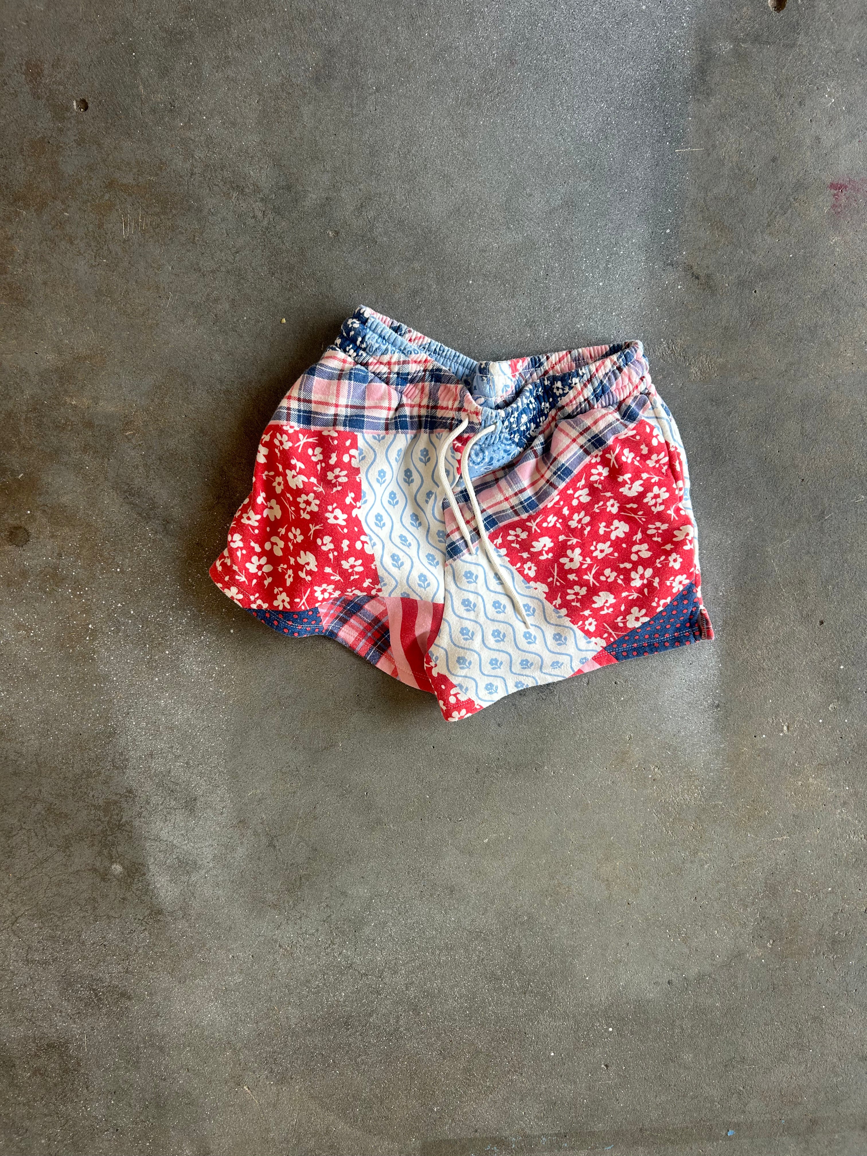 A pair of patchwork shorts with red, white, and blue patterns, laid flat on a gray concrete surface