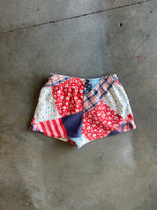 A pair of patchwork shorts made from red floral, blue polka dot, plaid, and striped fabrics, laid flat on a gray concrete floor