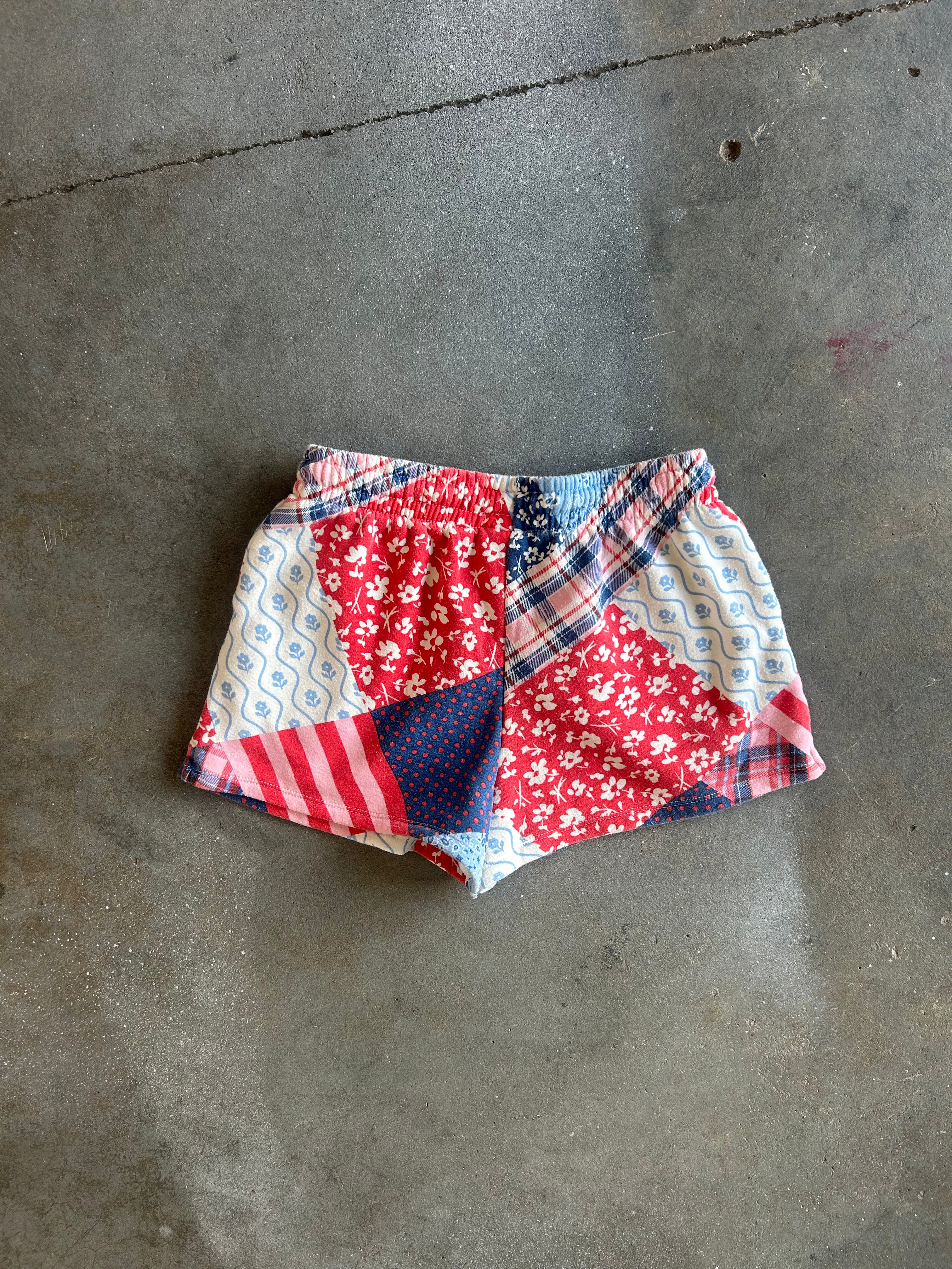 A pair of patchwork shorts made from red floral, blue polka dot, plaid, and striped fabrics, laid flat on a gray concrete floor