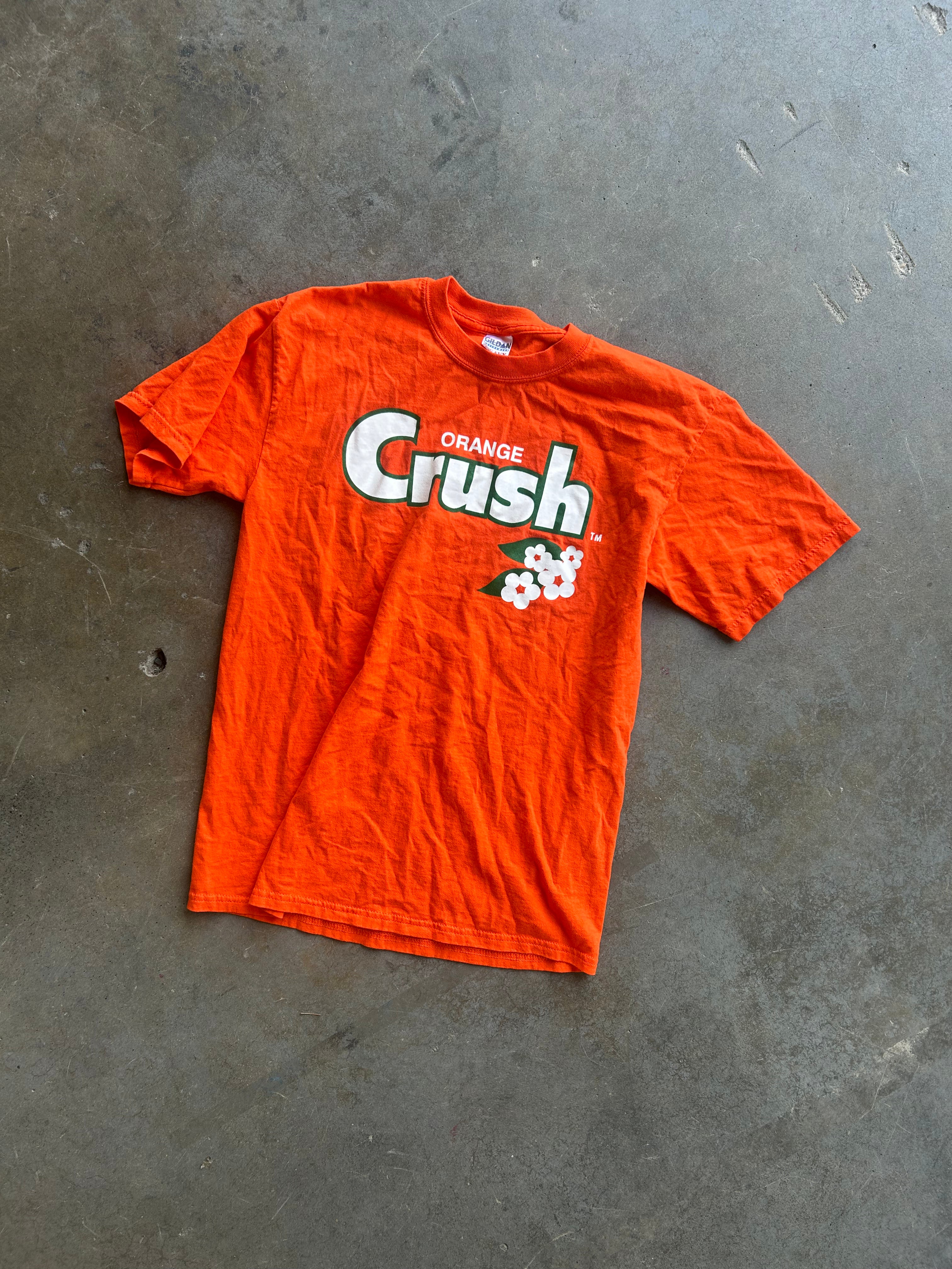 An orange short-sleeve graphic t-shirt with 'ORANGE Crush' printed in white and black on the front, laid flat on a textured gray concrete floor