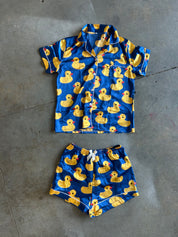 A two-piece pajama set laid flat on a gray concrete floor, featuring a short-sleeve button-front shirt and matching shorts in bright blue with an all-over yellow rubber duck print and white piping