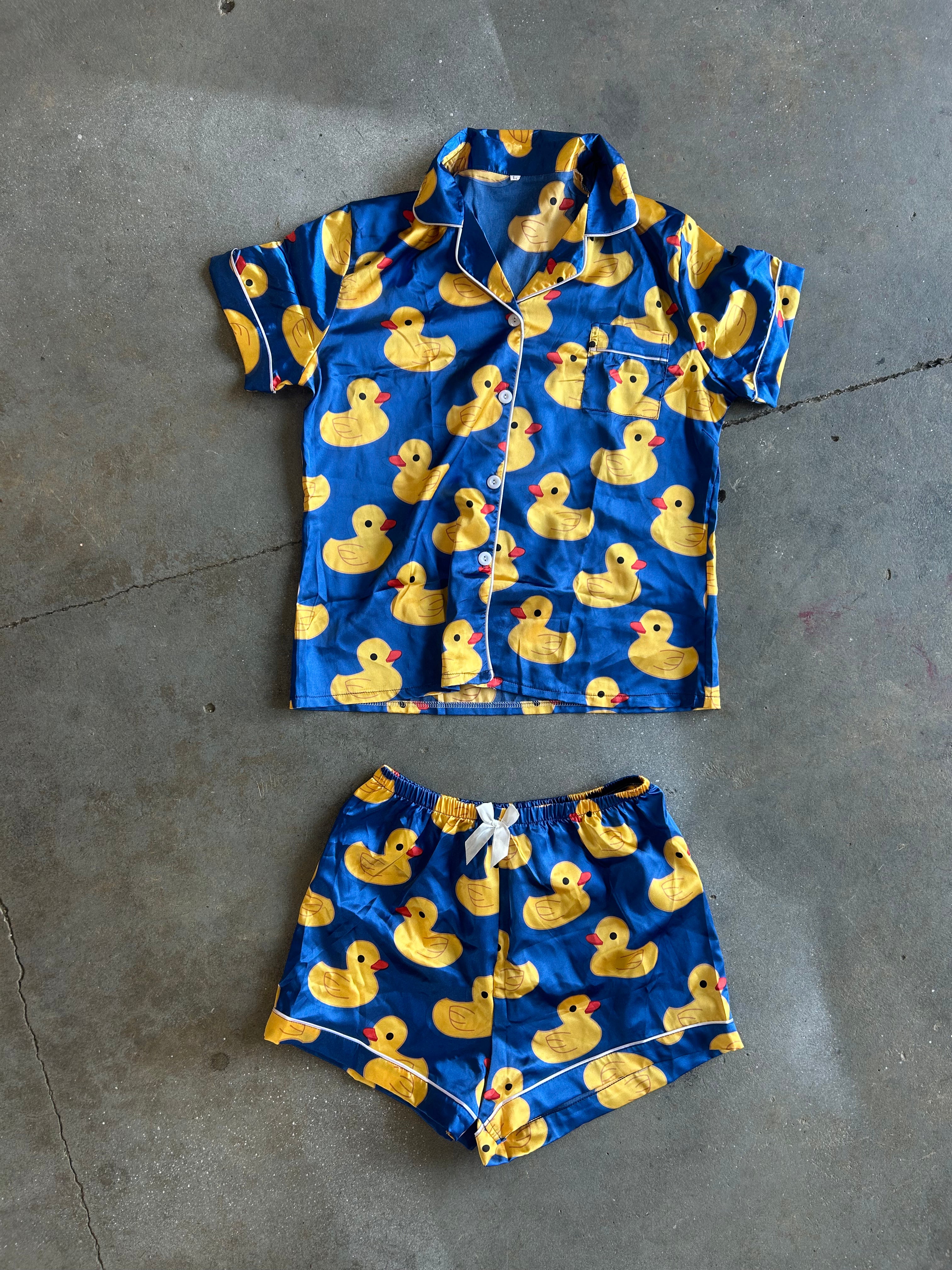 A two-piece pajama set laid flat on a gray concrete floor, featuring a short-sleeve button-front shirt and matching shorts in bright blue with an all-over yellow rubber duck print and white piping