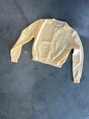 A pale yellow knit cardigan sweater with a button-up front, laid flat on a textured dark gray concrete floor