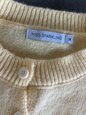 Close-up of a cream-colored knit cardigan sweater showing the ribbed collar, a white button, and a white fabric label that reads 'MISS SPARKLING' with size 'M'