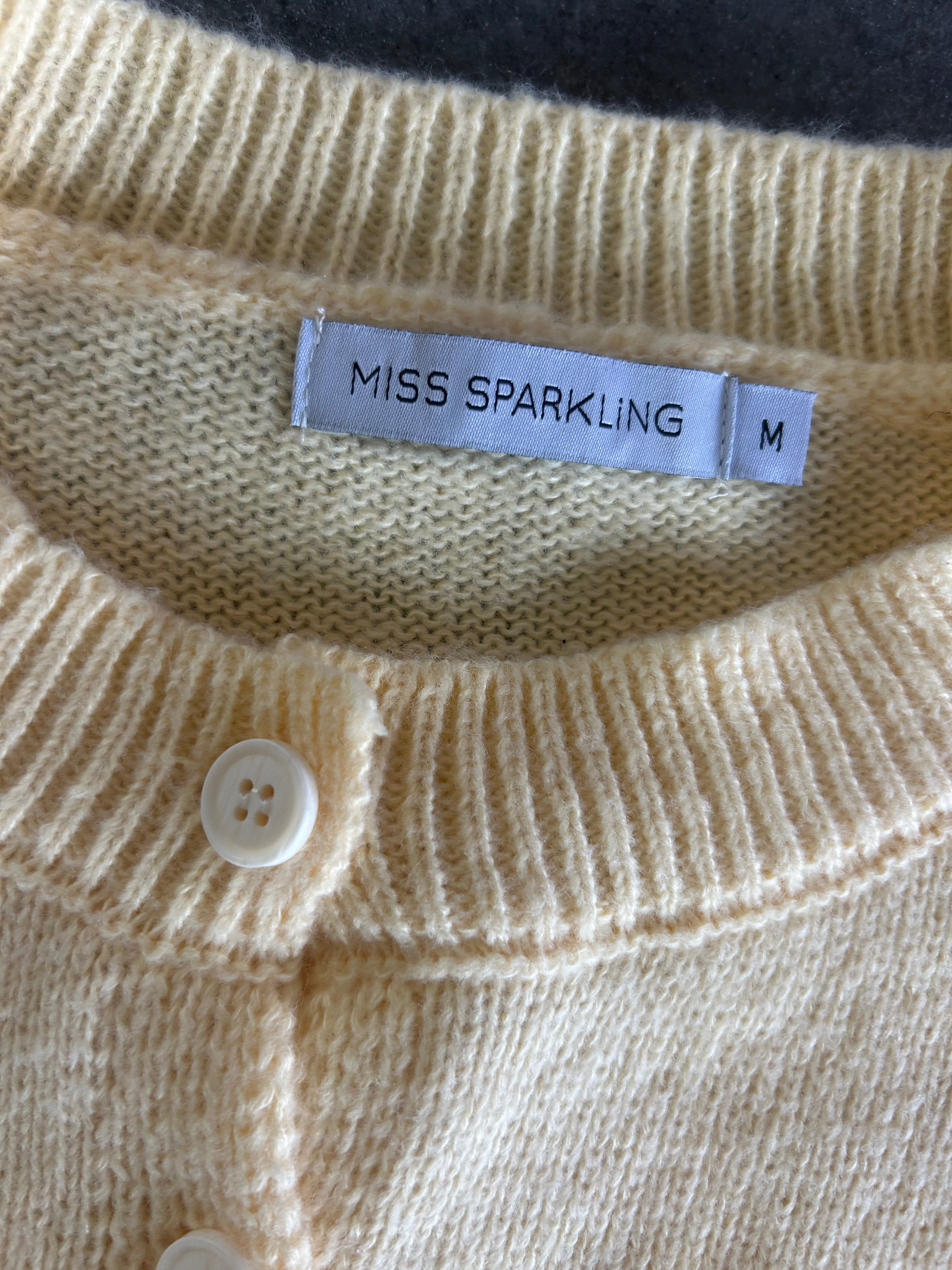 Close-up of a cream-colored knit cardigan sweater showing the ribbed collar, a white button, and a white fabric label that reads 'MISS SPARKLING' with size 'M'