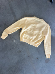 A pale yellow cropped knit sweater with long sleeves and ribbed cuffs and hem, laid flat on a dark gray concrete floor