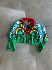 A cropped knit zip-up cardigan with a colorful cartoon landscape design, featuring rainbows, clouds, flowers, and smiling faces on a turquoise and green background, laid flat on a gray concrete floor